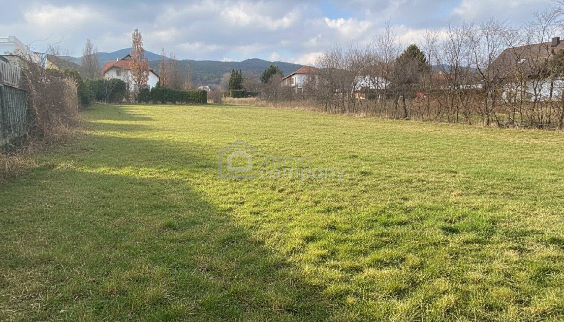 Weitläufiges grünes Grundstück unter bewölktem Himmel, mit Häusern und Bergen im Hintergrund.