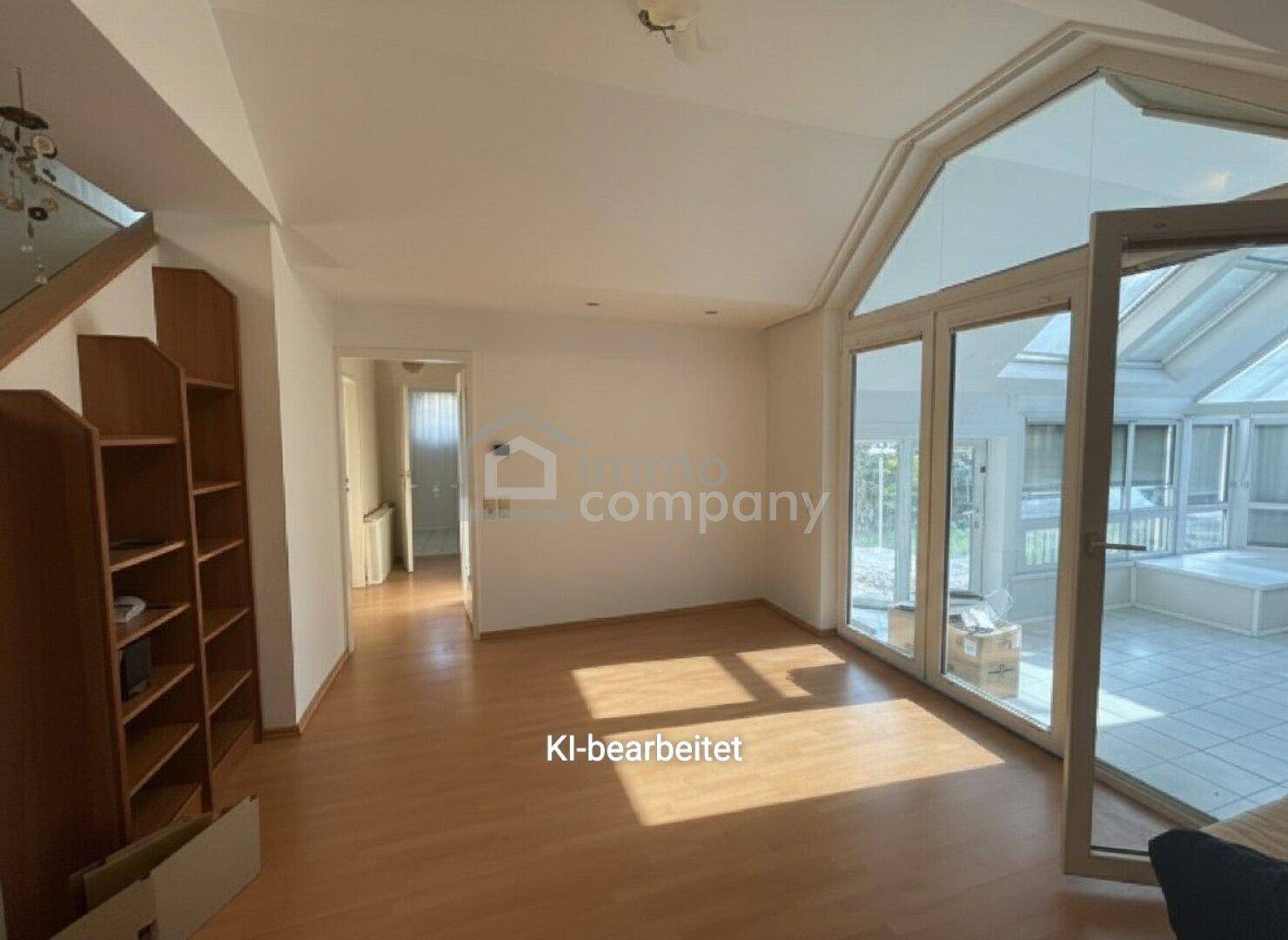 Leerer Wohnbereich mit hellem Parkettboden und großen Fenstern, die zum Wintergarten führen.