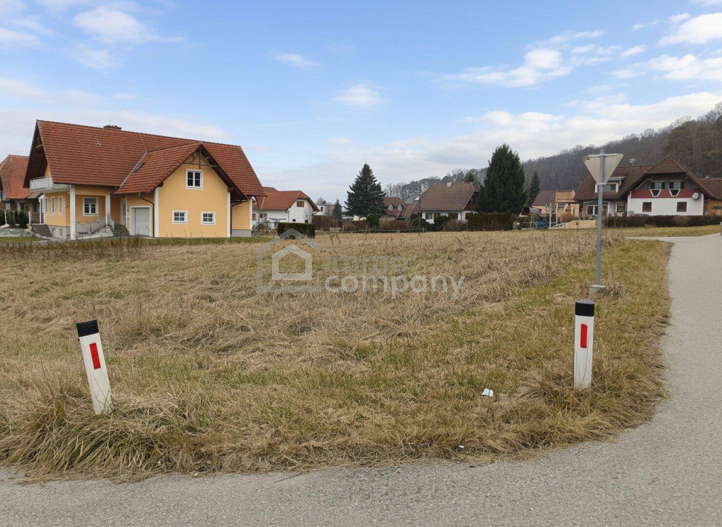 Unbebautes Grundstück mit einem gelben Haus im Vordergrund und einer hügeligen, bewaldeten Landschaft im Hintergrund.
