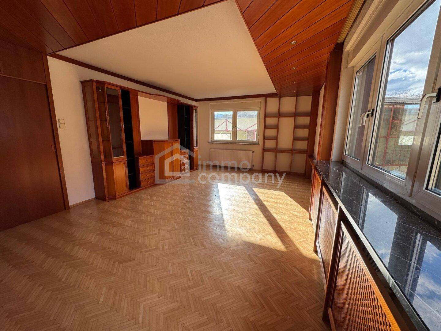 Leeres Wohnzimmer mit Parkettboden, Holzdecke und großen Fenstern.