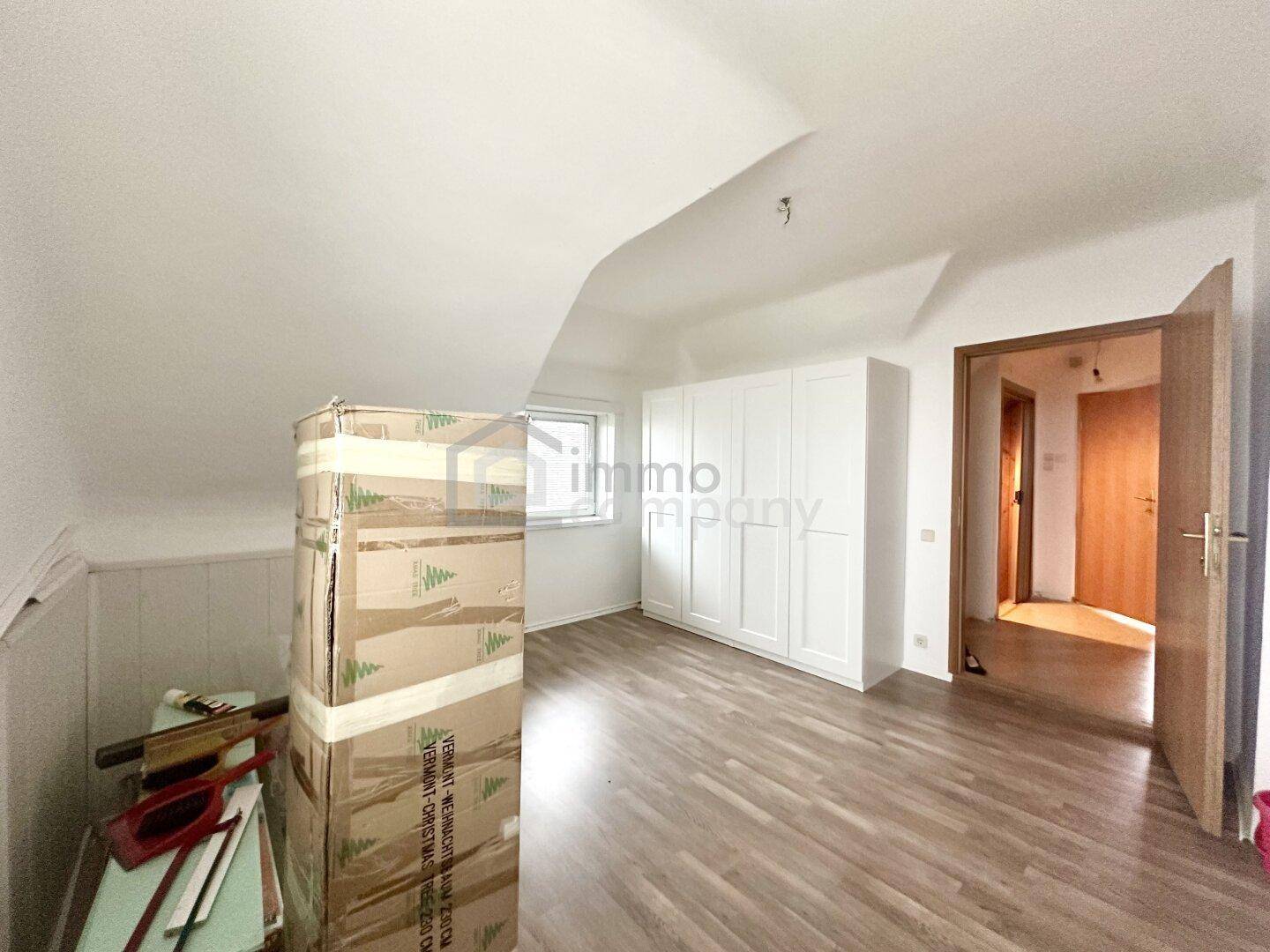 Dachzimmer mit Laminatboden, weißem Kleiderschrank und Zugang zu einem weiteren Raum.