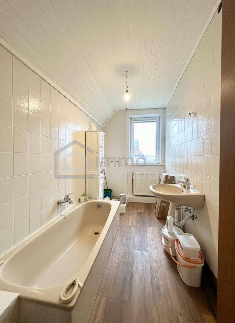 Badezimmer mit Badewanne, Waschbecken, Fenster und weißen Fliesen an den Wänden.