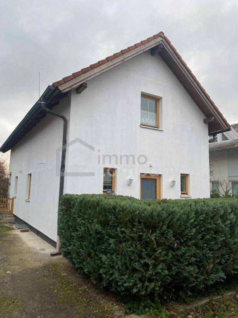 Seitenansicht des Hauses mit weißer Fassade, Satteldach und einer grünen, gepflegten Hecke.