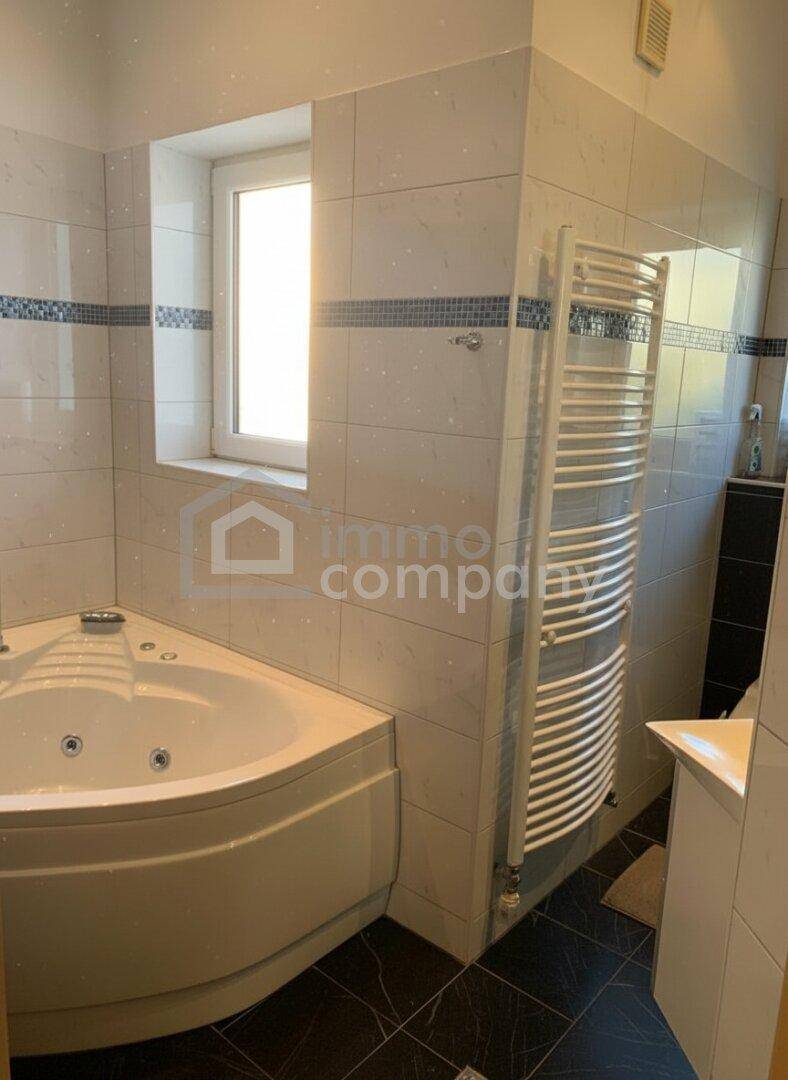 Helles Badezimmer mit Eckbadewanne, Fliesenboden und beheiztem Handtuchhalter am Fenster.