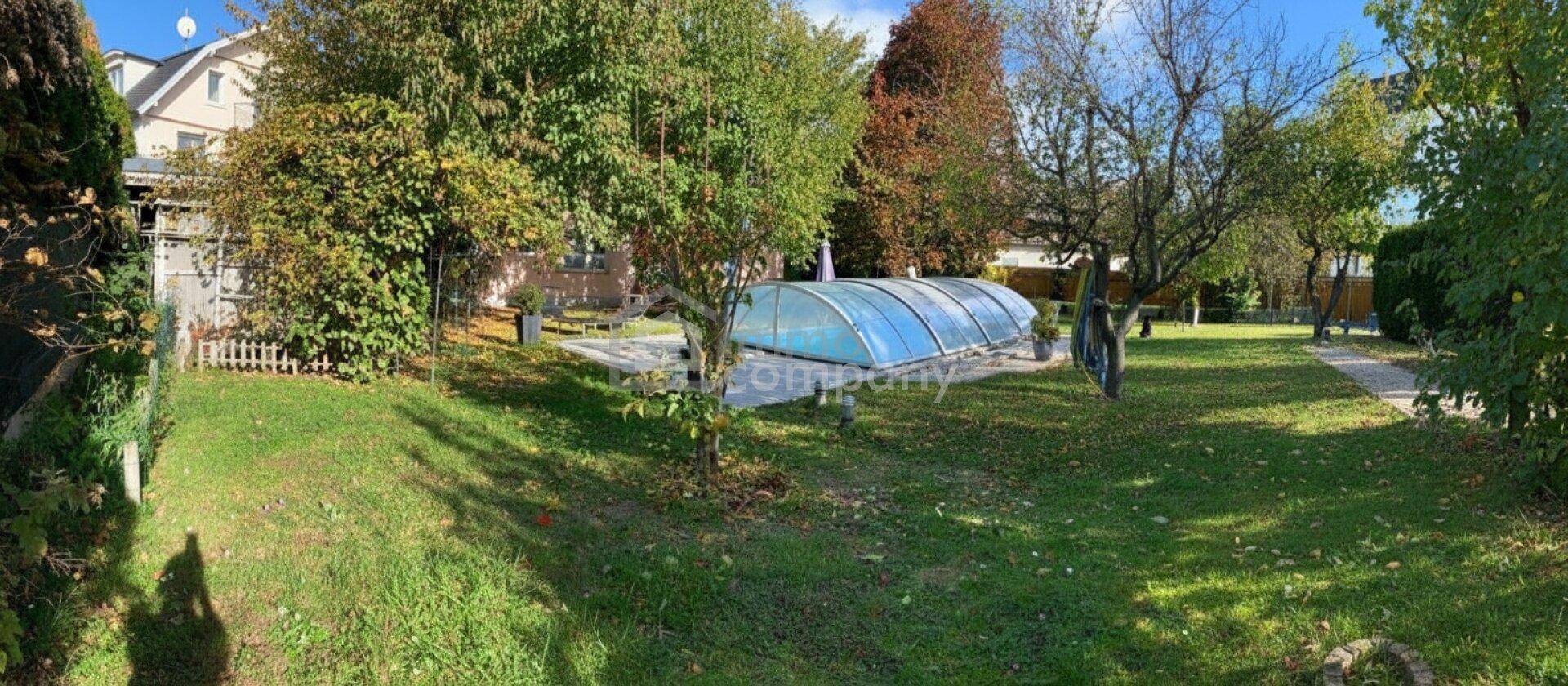 Großer grüner Garten mit Rasenfläche, Bäumen und einem überdachten Swimmingpool im Hintergrund.
