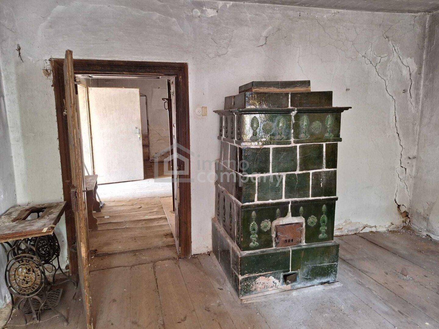 Rustikaler Raum mit grünem Kachelofen, Holzdielenboden und sichtbaren Wandrissen.