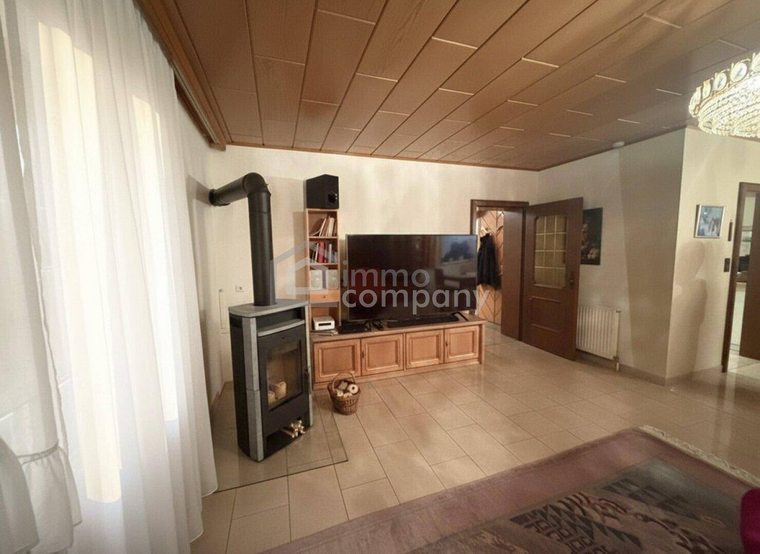 Geräumiges Wohnzimmer mit Kaminofen, Fernseher und Zugang zu weiteren Räumen.