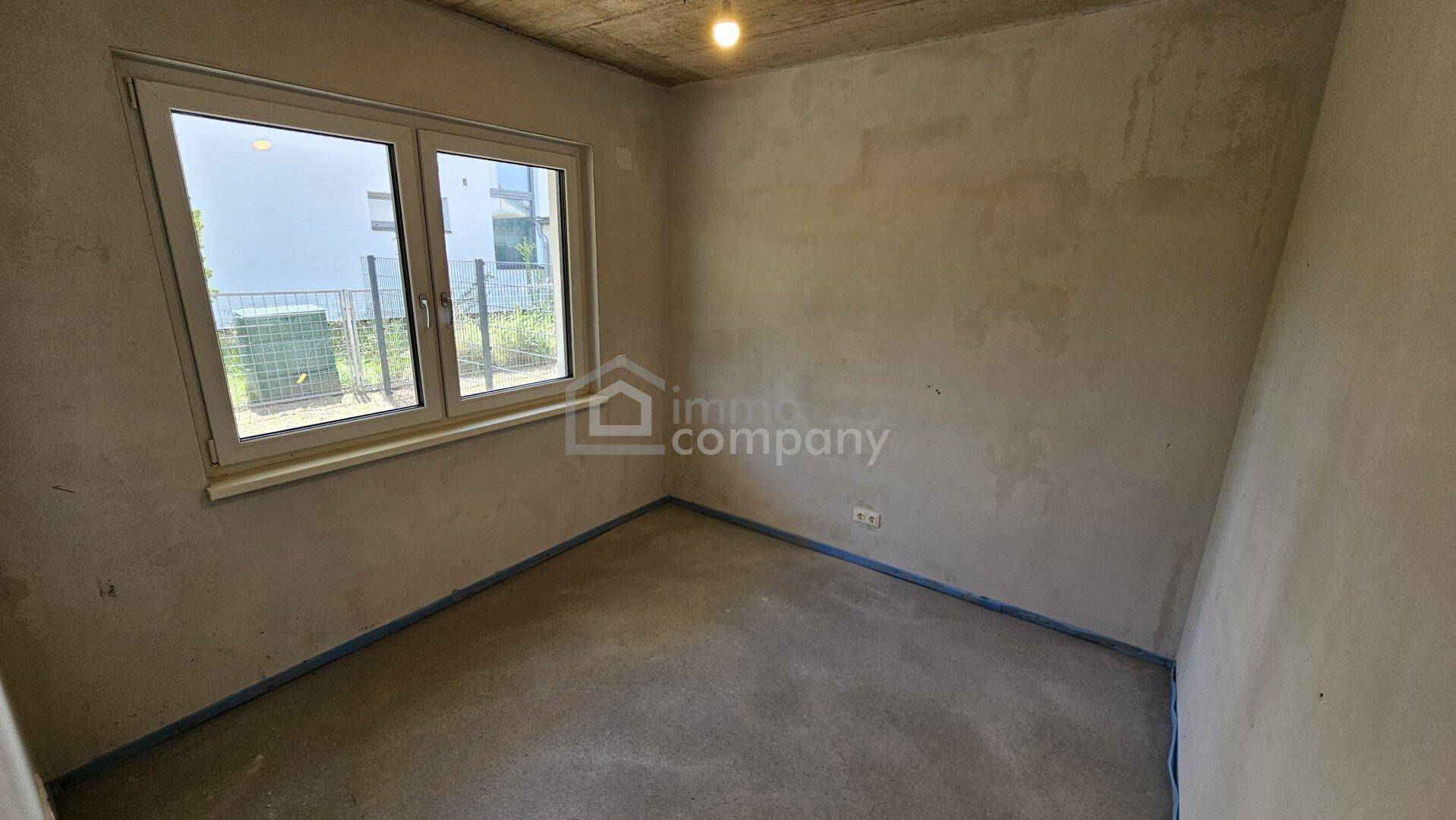 Unfertiges Zimmer im Rohbau mit Fenster, Betonoptik an der Decke und Blick ins Grüne.