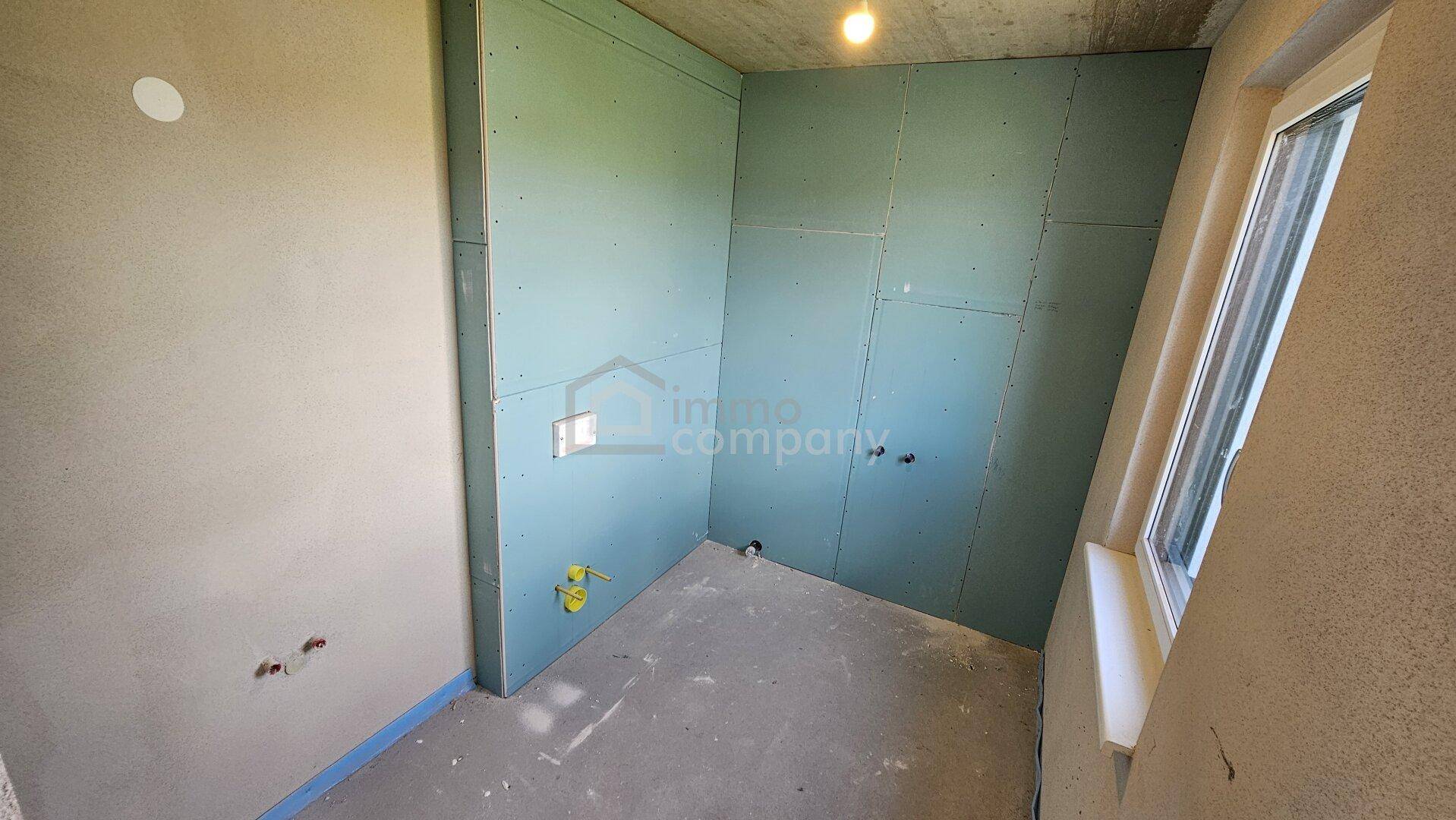 Unfertiges Badezimmer im Rohbau mit Fenster und vorbereiteten Installationen für Sanitäranlagen.