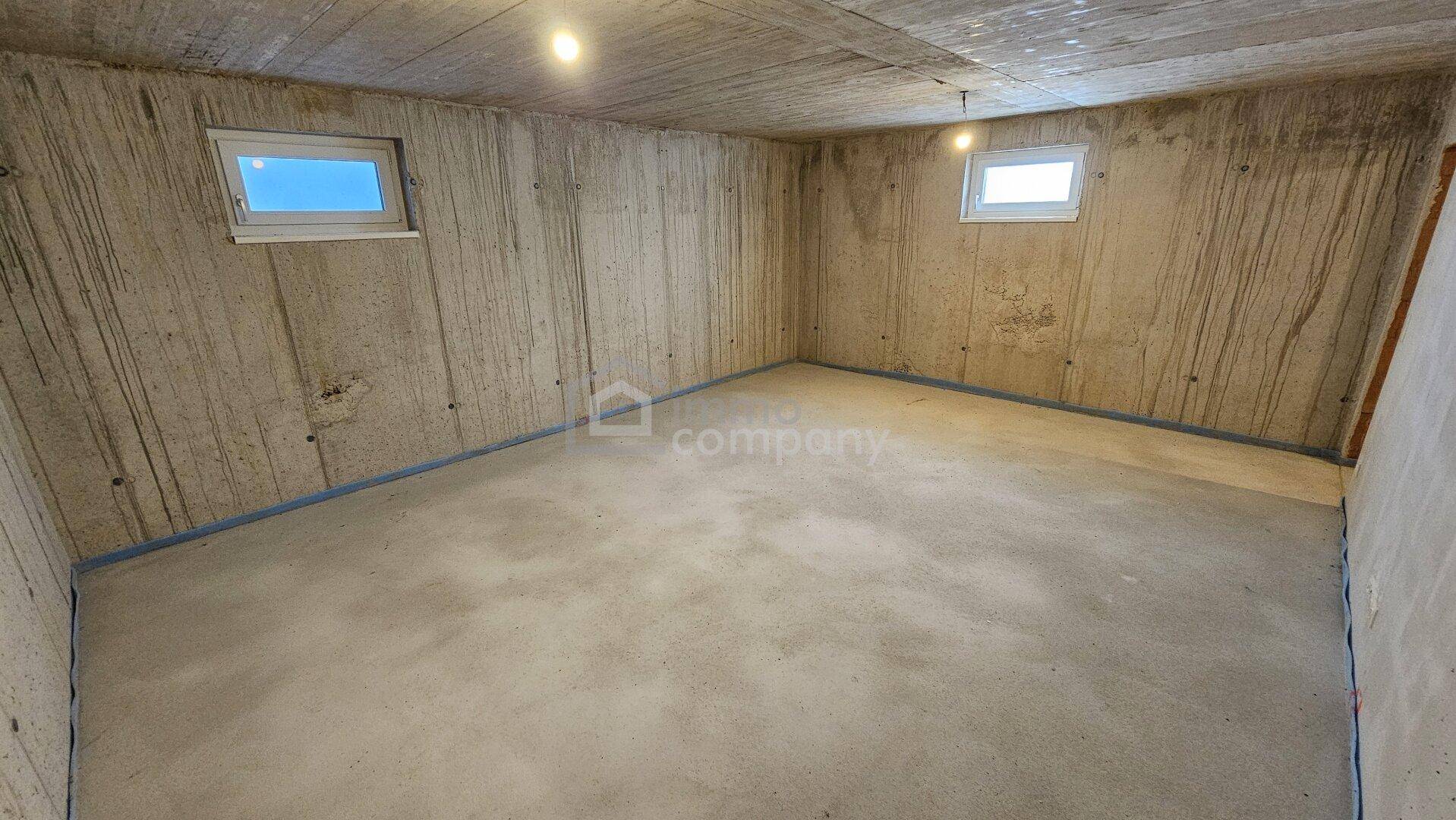 Kellerraum im Rohbau mit Betonwänden und zwei kleinen Fenstern, ideal als Lagerfläche.
