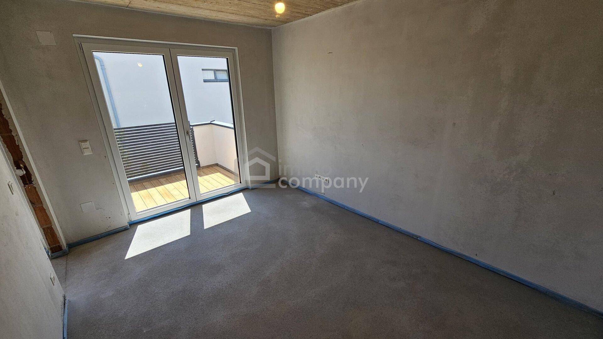 Unfertiges Zimmer im Rohbau mit Zugang zu einem Balkon und Betonoptik an der Decke.