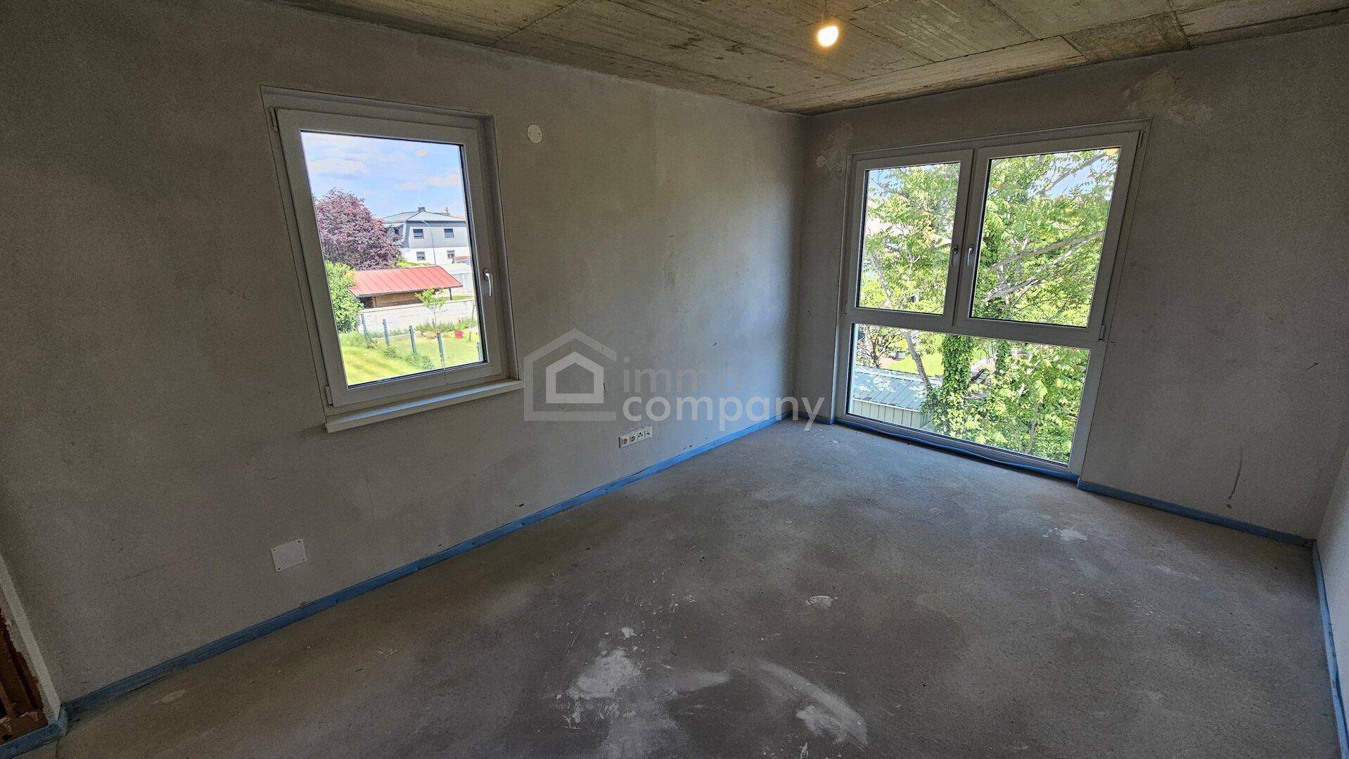 Geräumiges Zimmer im Rohbau mit zwei Fenstern, Betonoptik an der Decke und Blick ins Grüne.