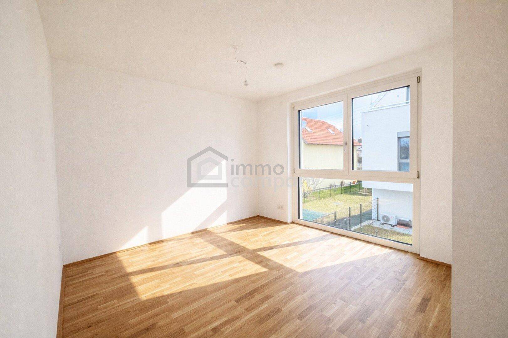 Lichtdurchflutetes, leeres Zimmer mit großem Fenster und Blick auf den Gartenbereich.