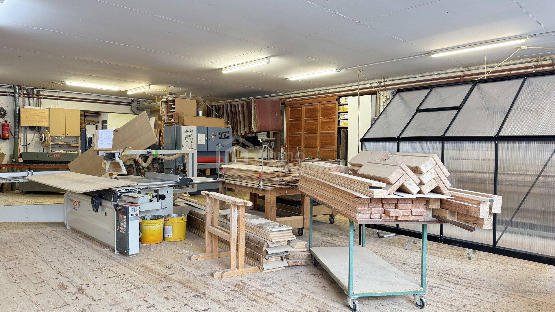 Helle Holzwerkstatt mit großen Fenstern, Maschinen und gestapeltem Holz für die Verarbeitung.