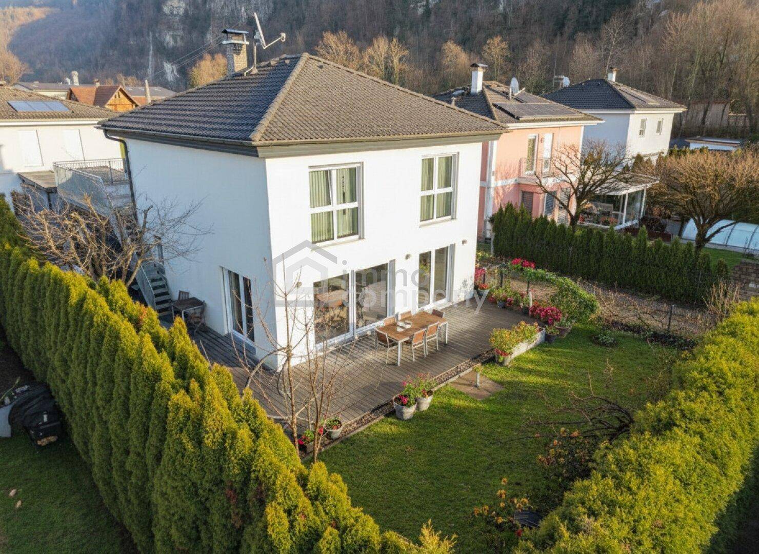 Drohnenaufnahme eines modernen Einfamilienhauses mit großer Terrasse, gepflegtem Garten und umliegender Hecke.