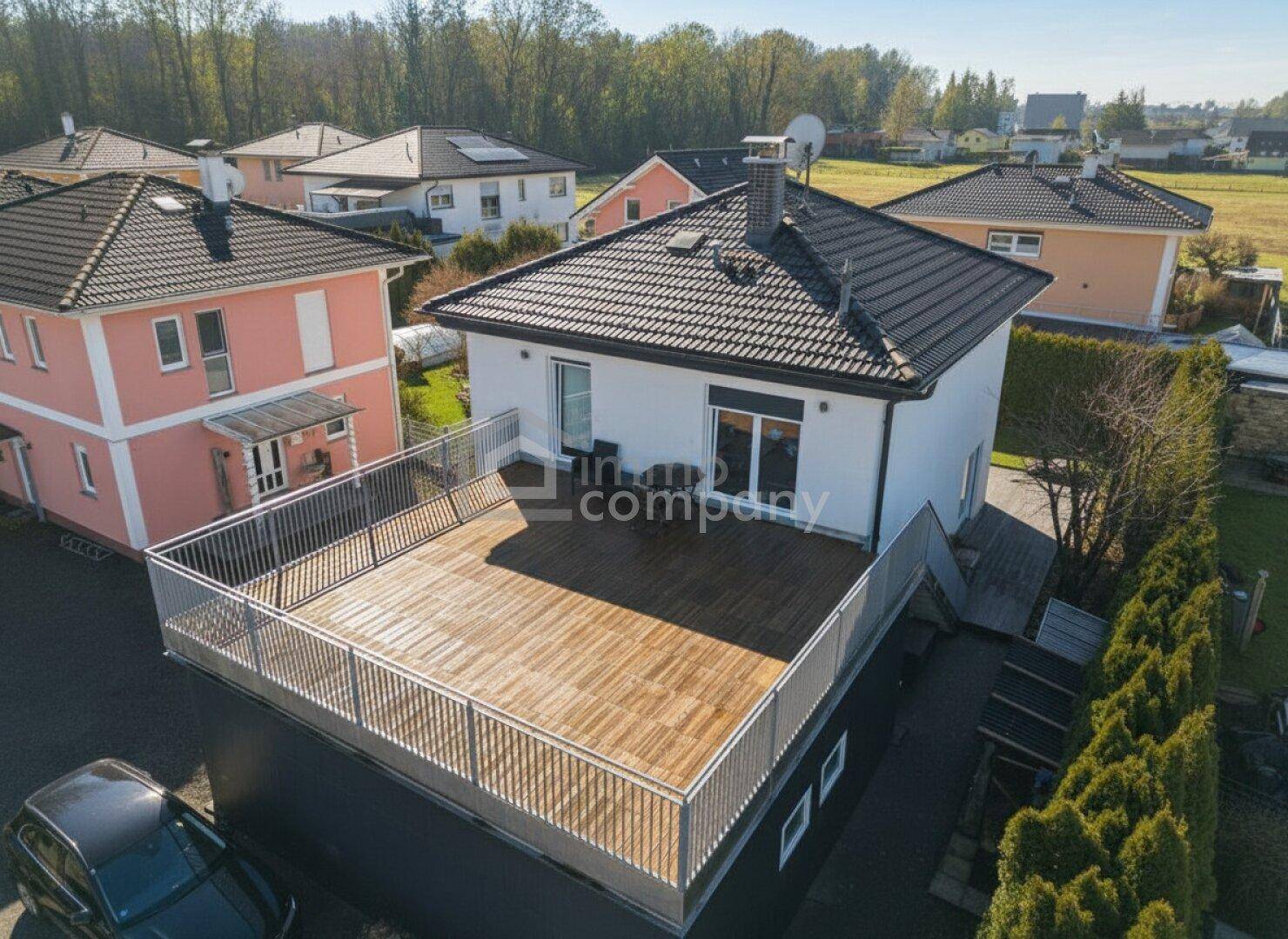 Luftaufnahme eines Einfamilienhauses mit großer Holzterrasse im Obergeschoss und umliegender Hecke.