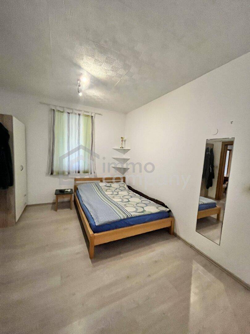 Helles Schlafzimmer mit einfachem Holzbett, Laminatboden und einem Fenster mit Blick nach draußen.