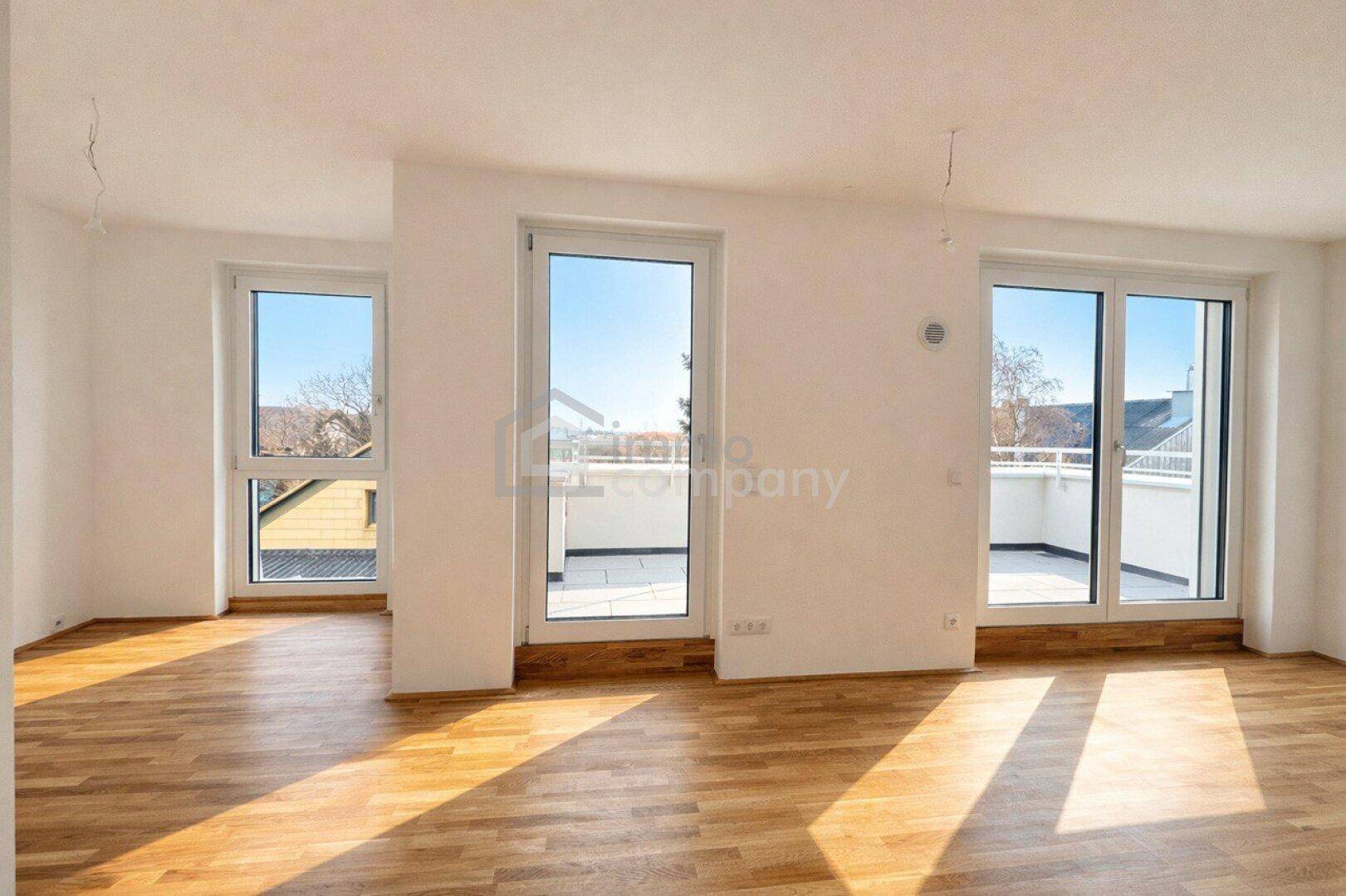 Heller, leerer Wohnraum mit großen Fenstern, Holzboden und Zugang zu einem Balkon.