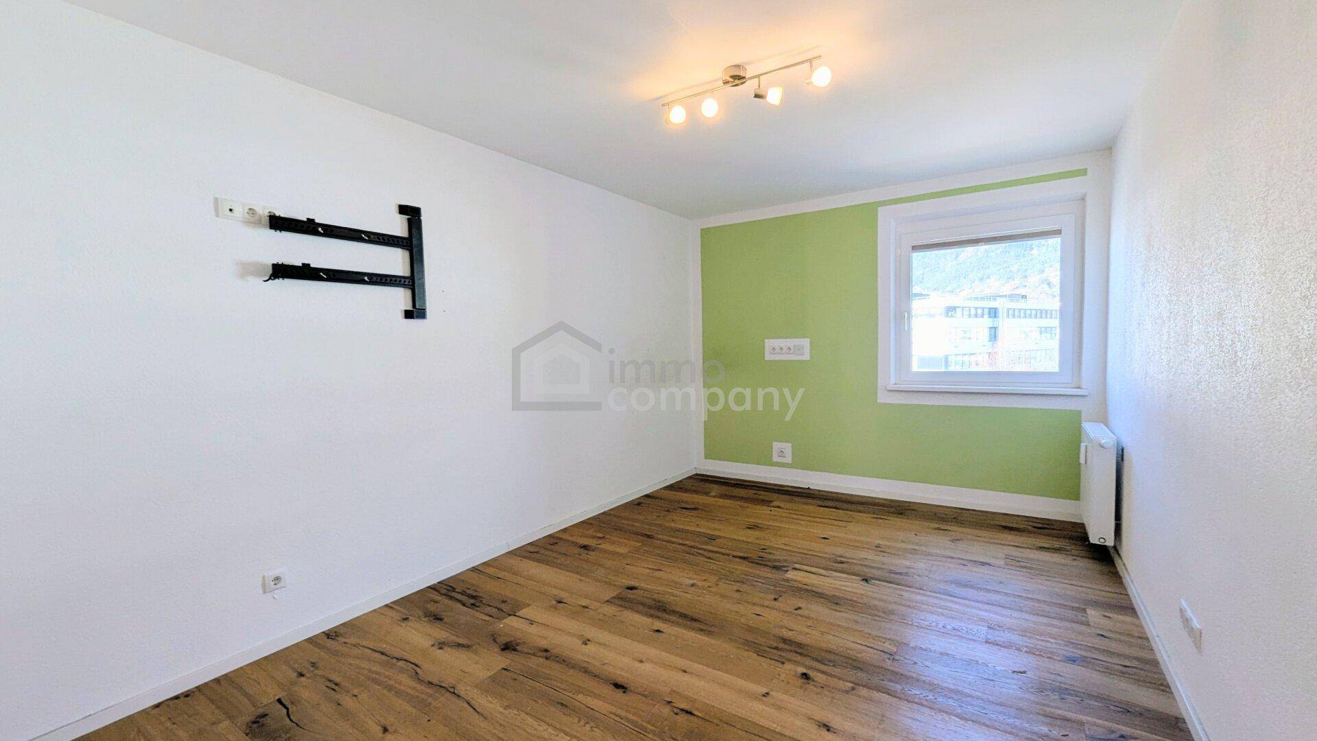 Helles Zimmer mit Holzboden, grüner Akzentwand und einem Fenster, vielseitig nutzbar.