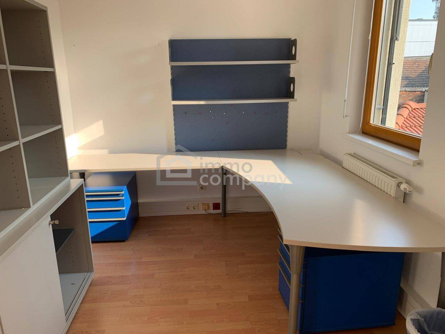 Funktioneller Bürobereich mit Eckschreibtisch, Regalen und blauen Akzenten, gut beleuchtet durch ein Fenster.