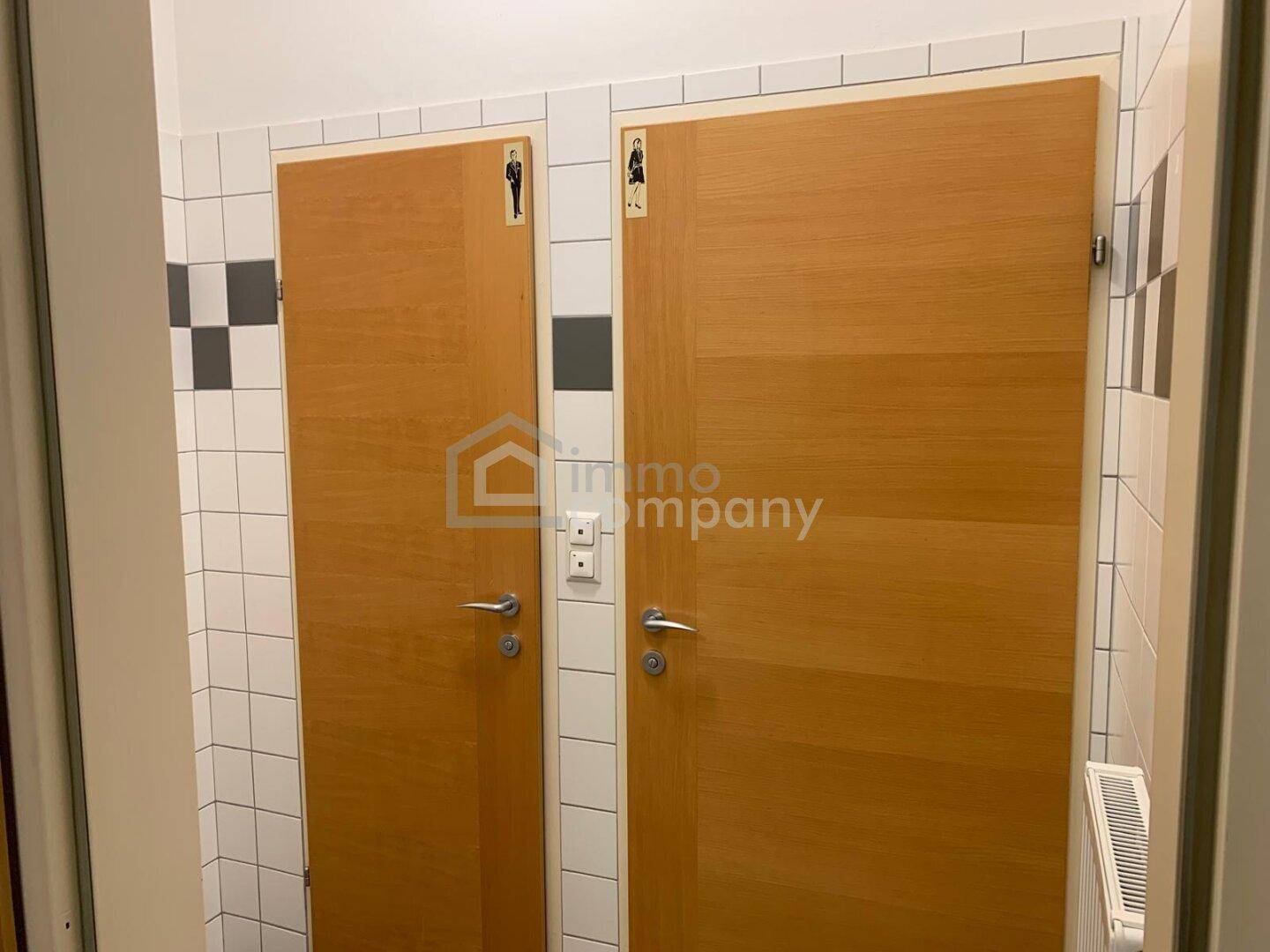 Zwei separate Toilettenkabinen mit Holztüren und weißen Fliesen, gekennzeichnet für Damen und Herren.