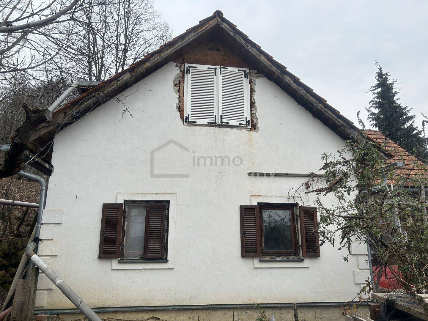 Rustikale Hausfassade mit weißen Fensterläden und sichtbaren Spuren der Zeit, umgeben von Bäumen.
