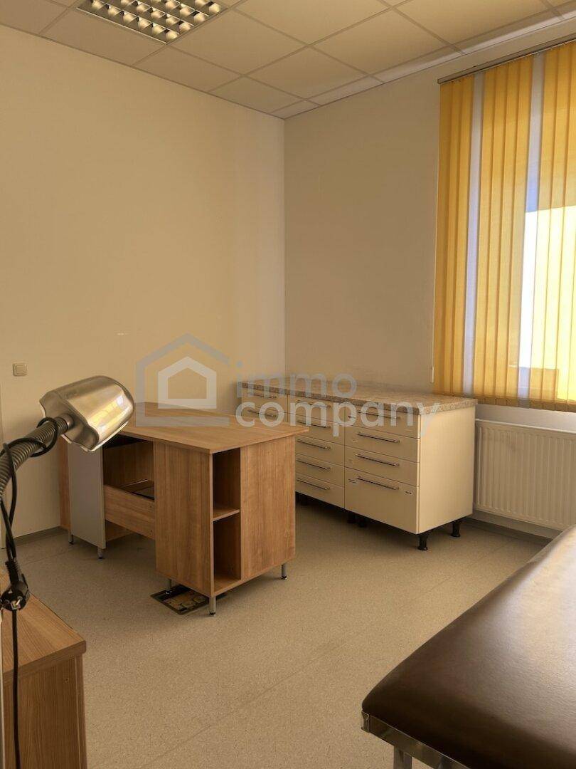 Geräumiges Büro oder Behandlungszimmer mit Schreibtisch, Kommode und Untersuchungsliege.