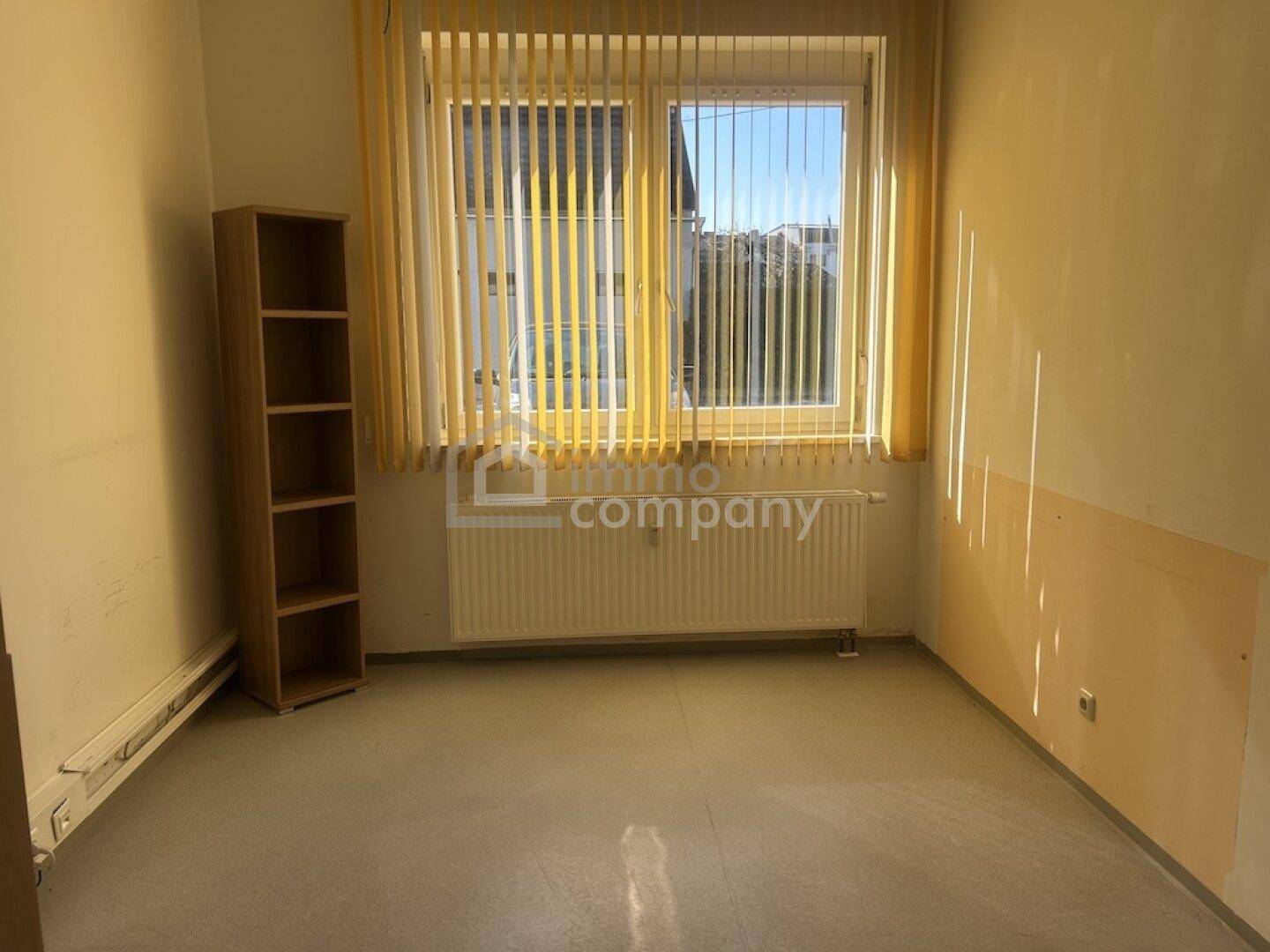 Leeres Zimmer mit großem Fenster, Jalousien und einem einfachen Holzregal.