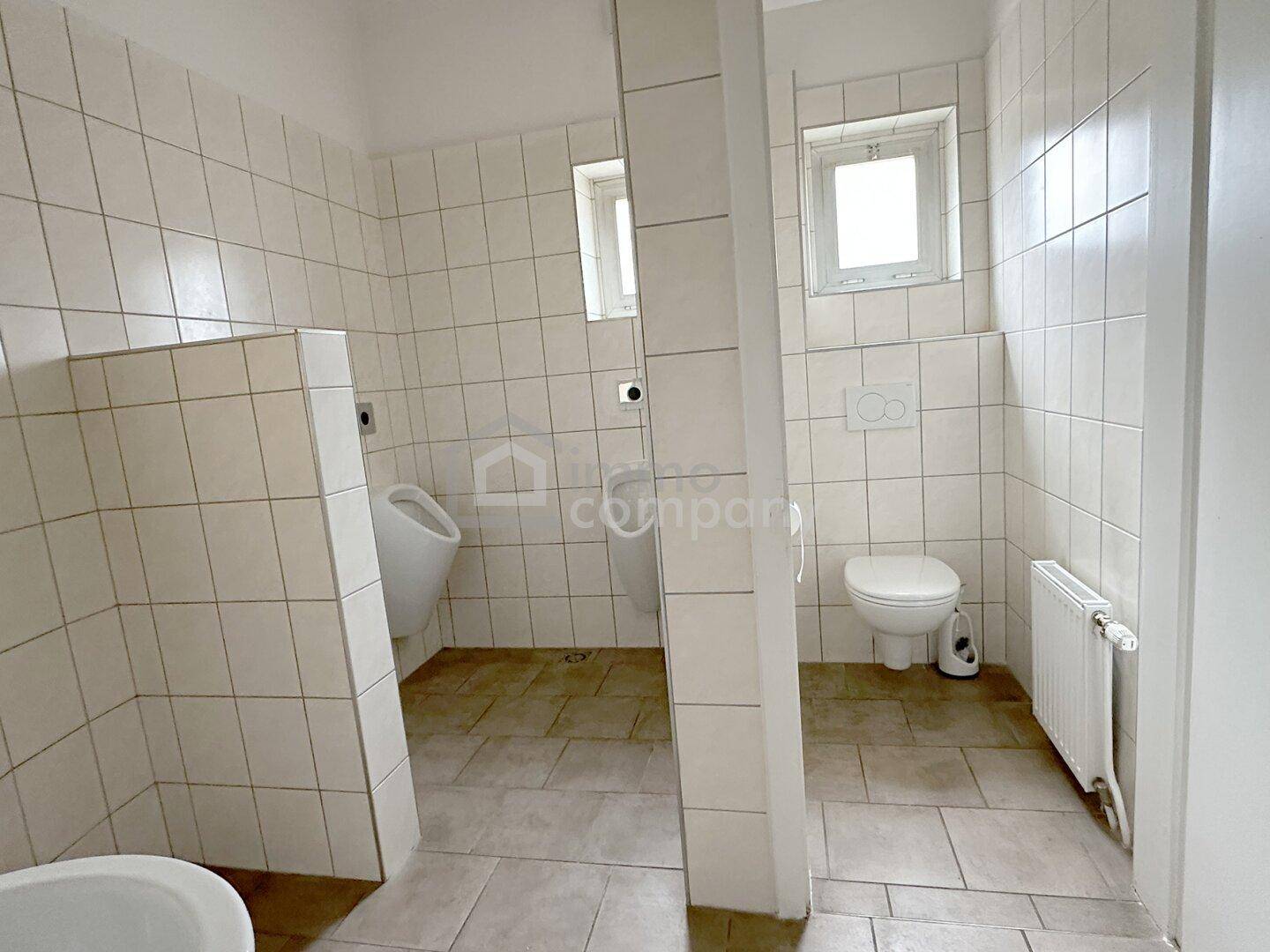 Gefliester Sanitärbereich mit Urinal, Toilette und Fenster, funktional und sauber.