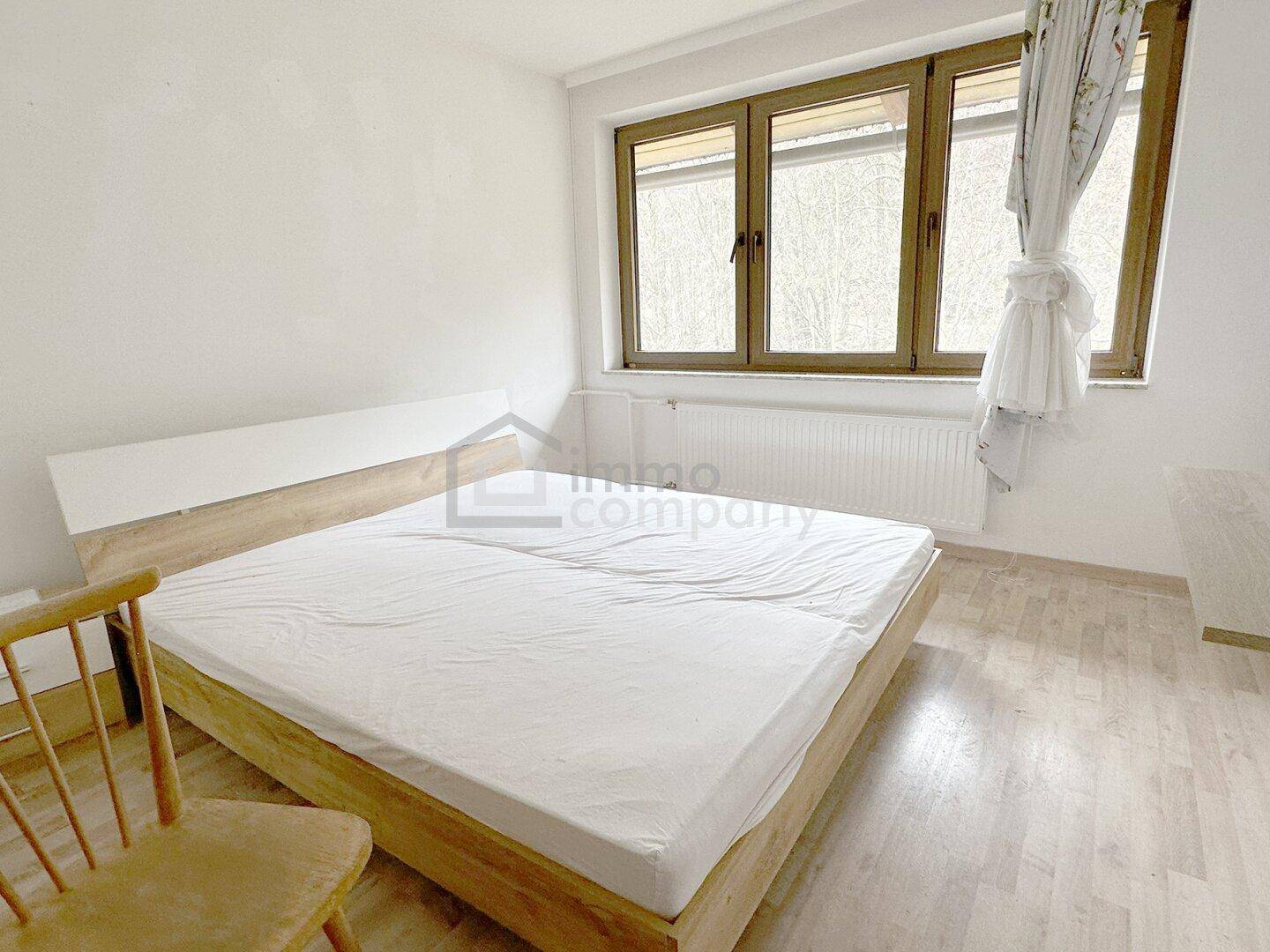 Helles Schlafzimmer mit Doppelbett, Laminatboden und großen Fenstern, schlicht eingerichtet.