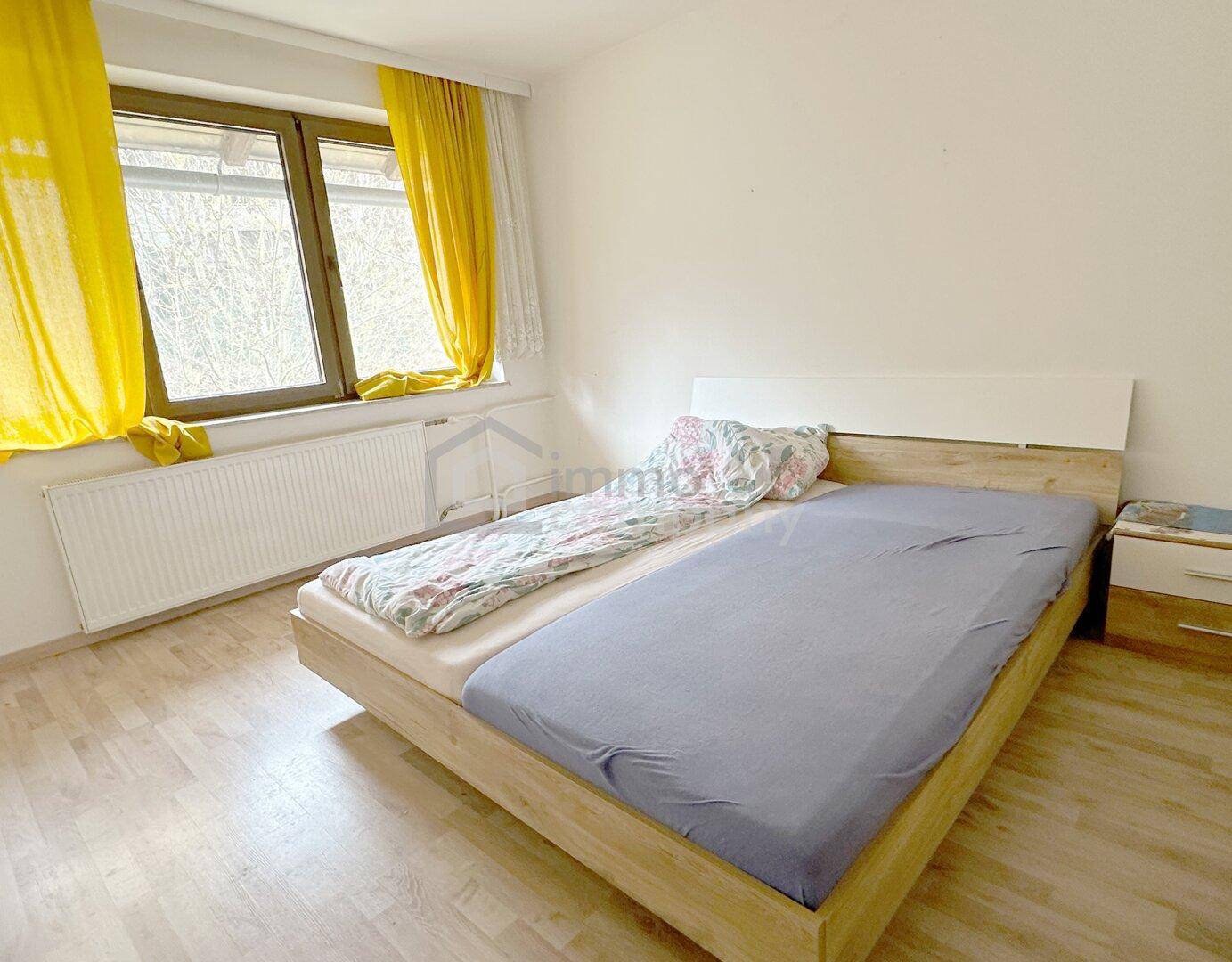 Helles Schlafzimmer mit Doppelbett, Laminatboden und gelben Vorhängen, freundlich gestaltet.