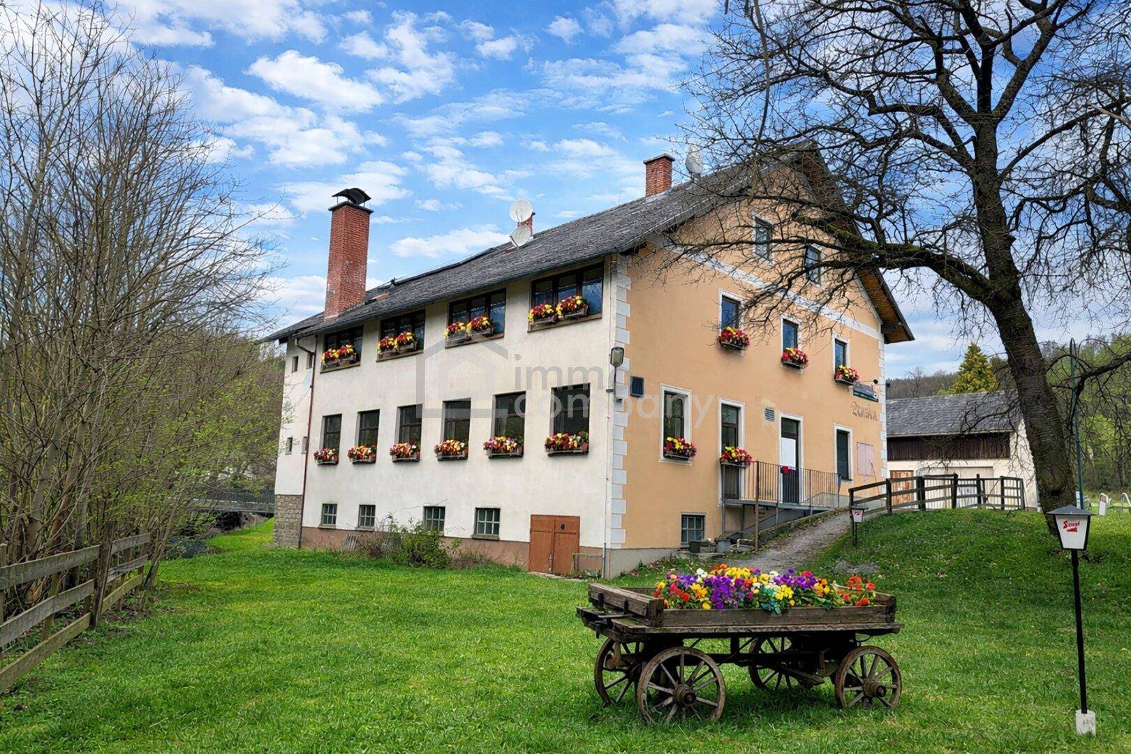 Historisches Gebäude mit blühenden Blumenkästen und einem dekorativen Blumenwagen im Vorgarten.