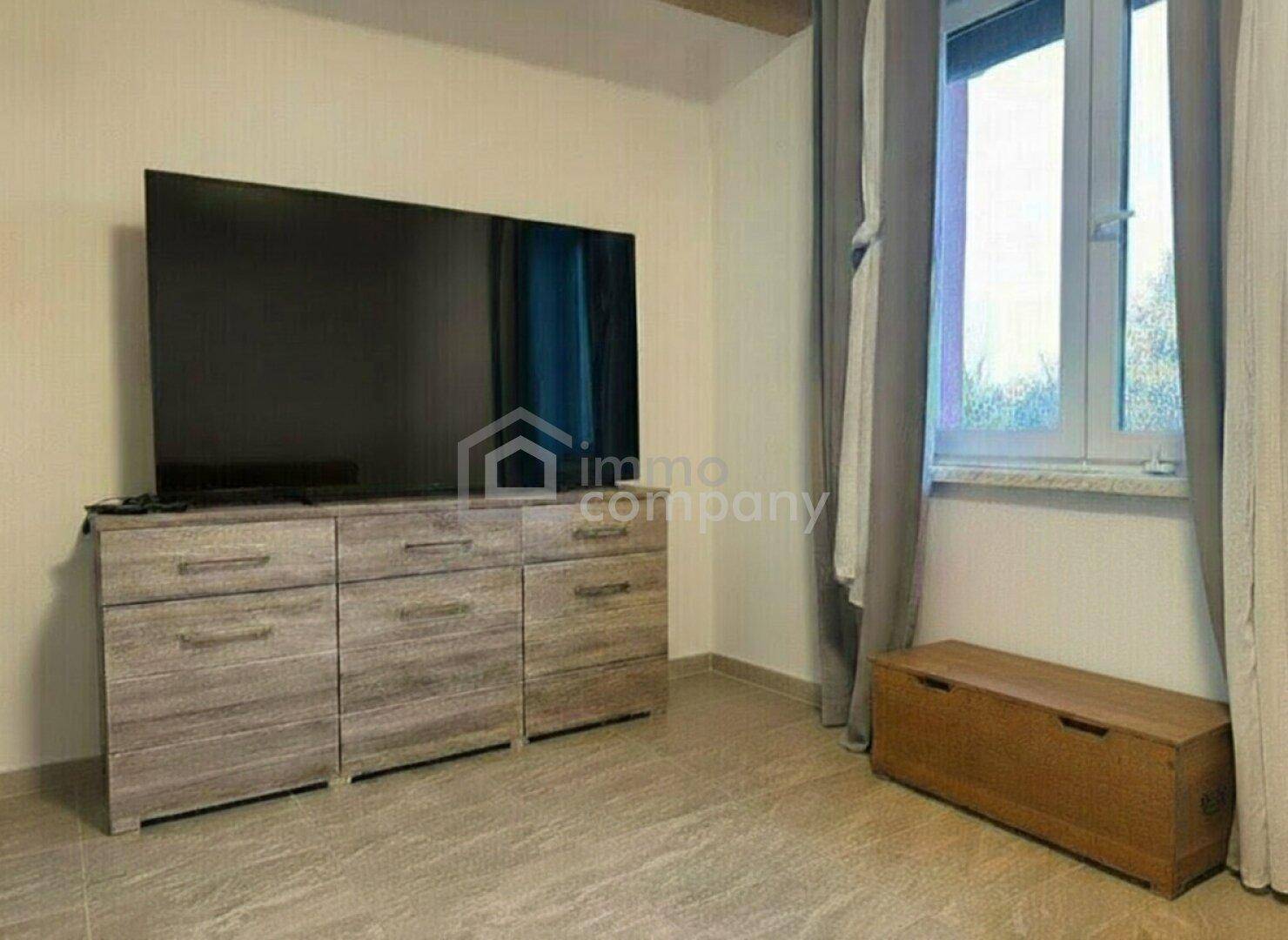 Raum mit großem Fernseher auf einer Kommode und Fenster mit Vorhängen.