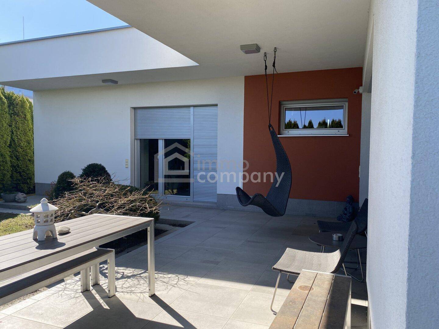 Überdachte Terrasse mit modernen Fliesen, Sitzgelegenheiten und einer Hängeschaukel im Gartenbereich.