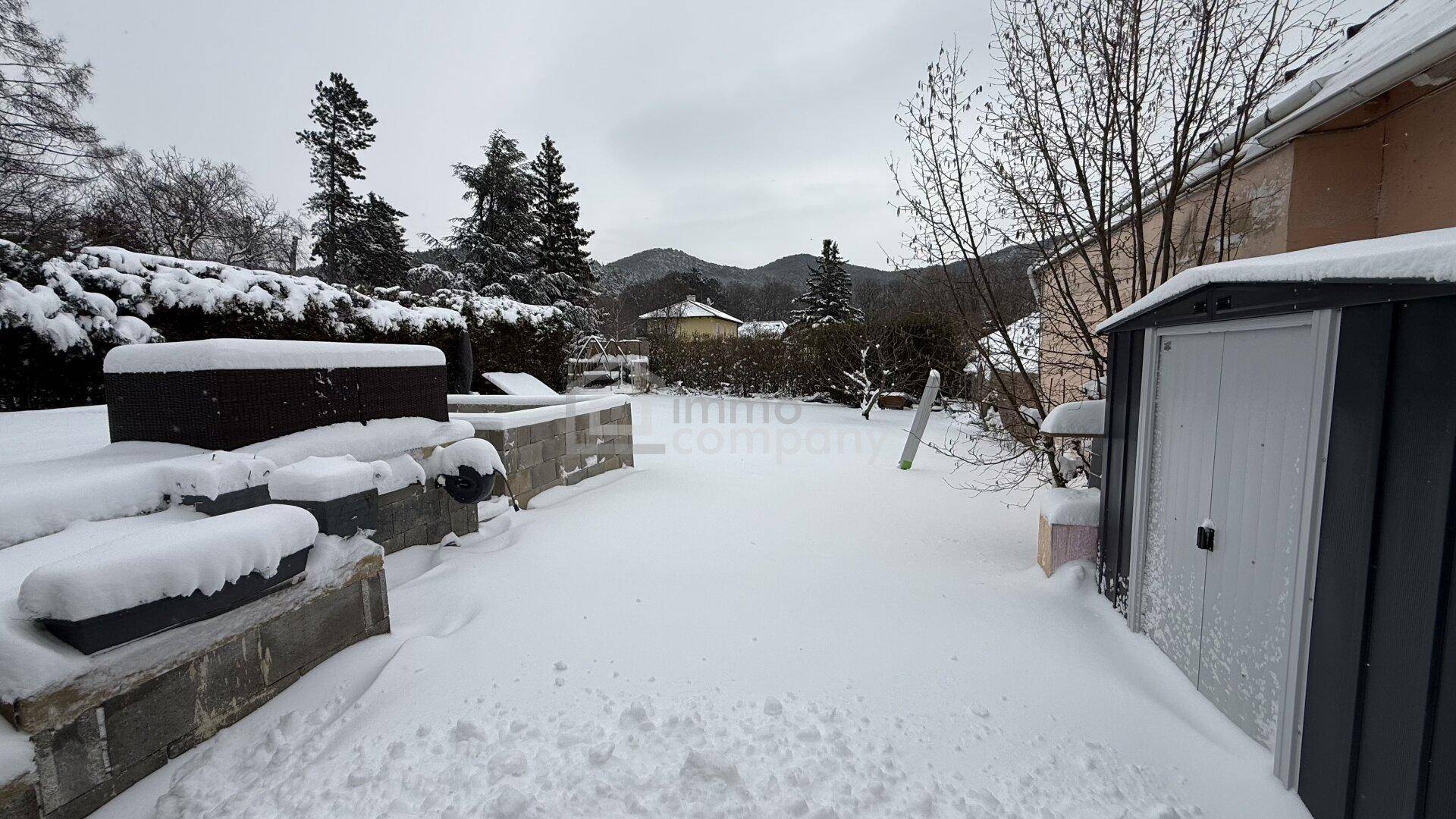Winterlicher Garten mit verschneiter Rasenfläche, Gartenhaus und Blick auf die umliegende Landschaft.