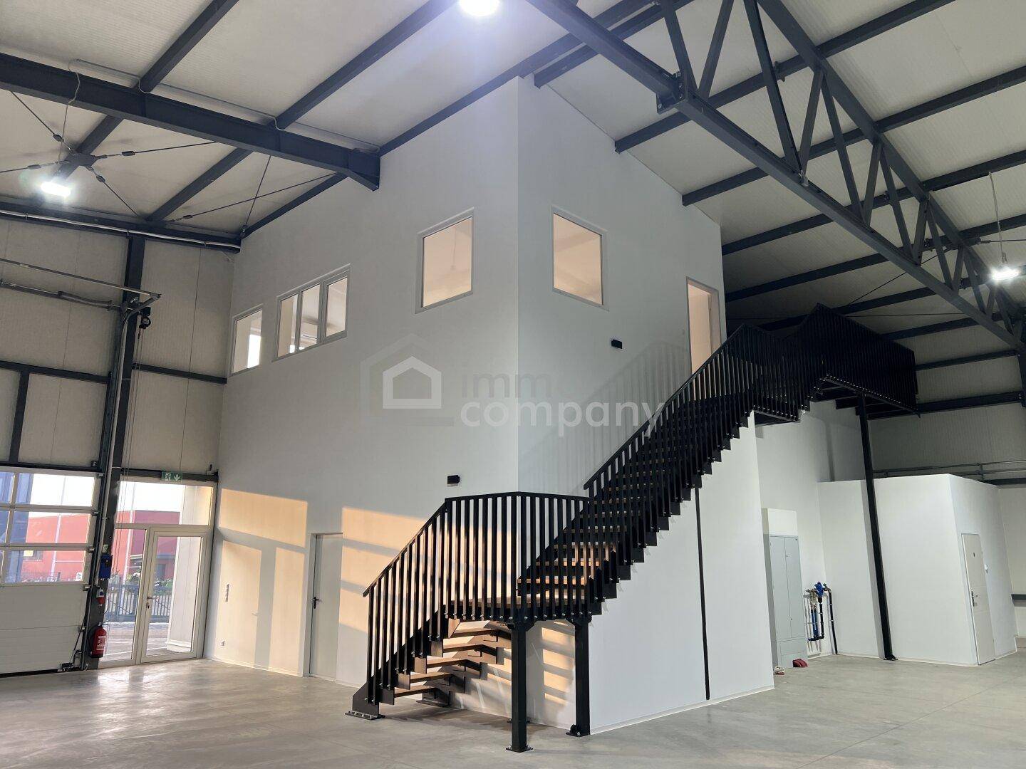 Geräumige Industriehalle mit hoher Decke, sichtbaren Stahlträgern und einer modernen Treppe zu einem Zwischengeschoss.