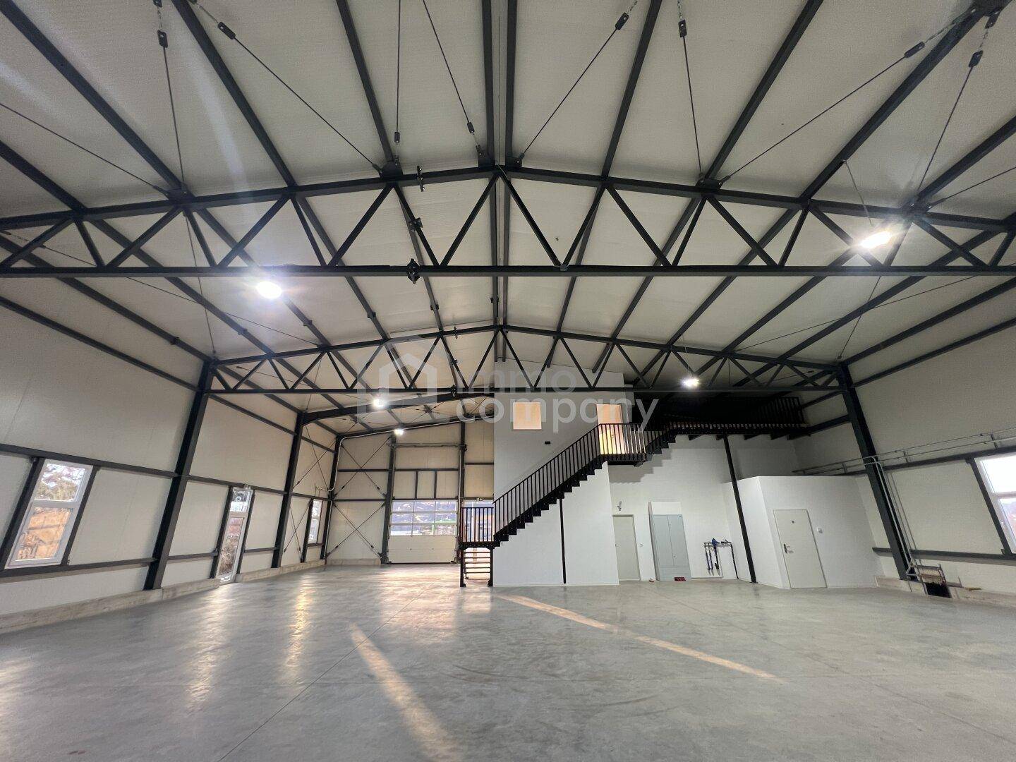 Große, helle Industriehalle mit hoher Decke, freiliegenden Stahlträgern und einem Zwischengeschoss mit Treppenzugang.