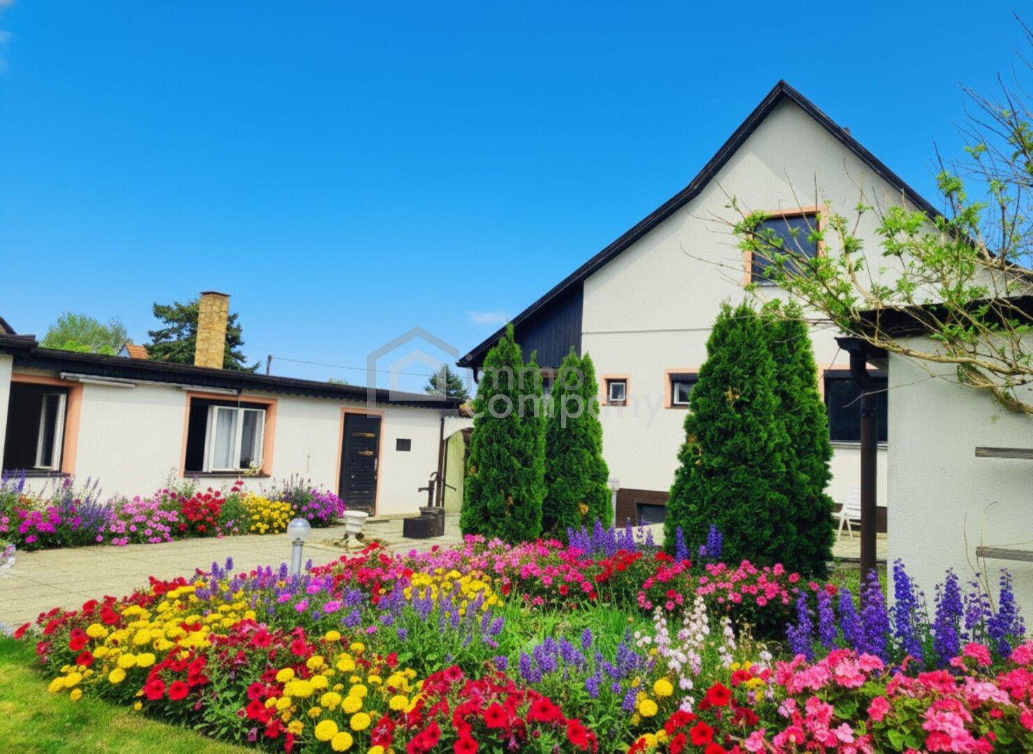 Hausfassade mit farbenfrohen Blumenbeeten und gepflegter Grünfläche im Vorgarten.