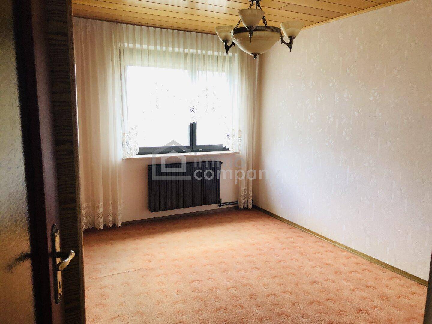 Heller Raum mit Teppichboden, großem Fenster und Heizkörper unter der Fensterbank.