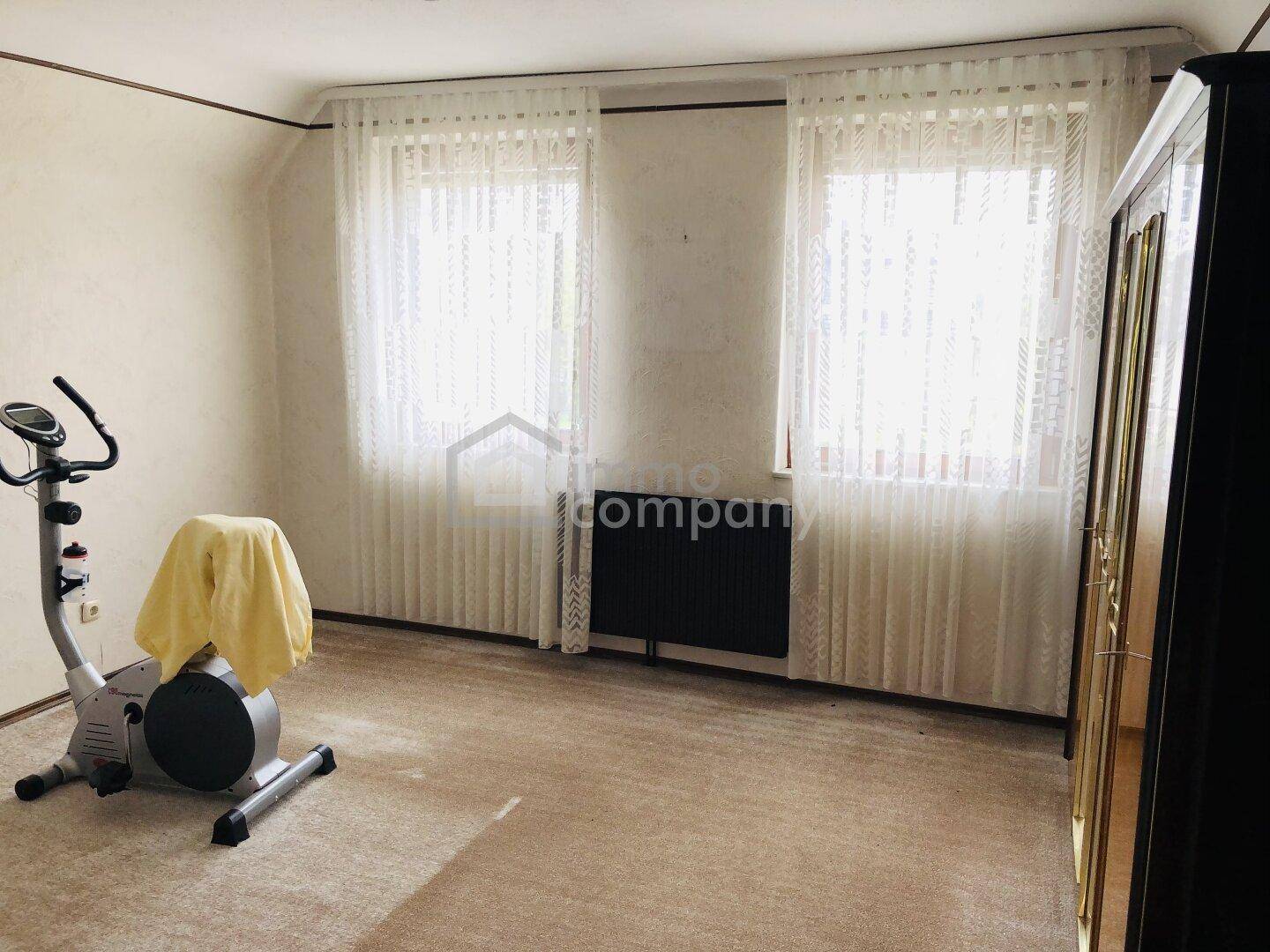 Heller Raum mit zwei Fenstern, Teppichboden und einem Heimtrainer.