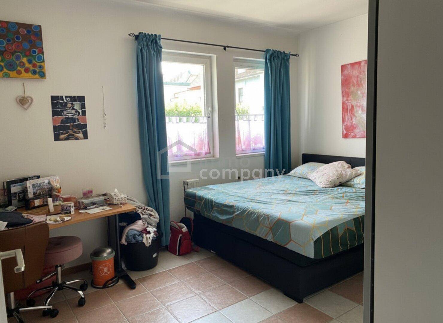Helles Schlafzimmer mit zwei Fenstern, Schreibtisch und farbenfroher Dekoration.