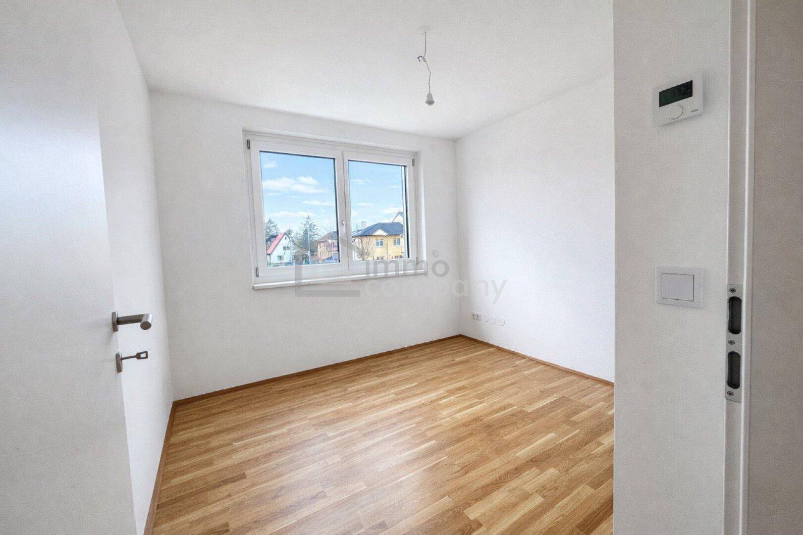 Helles Zimmer mit modernem Parkettboden, weißen Wänden und Blick ins Grüne durch das Fenster.