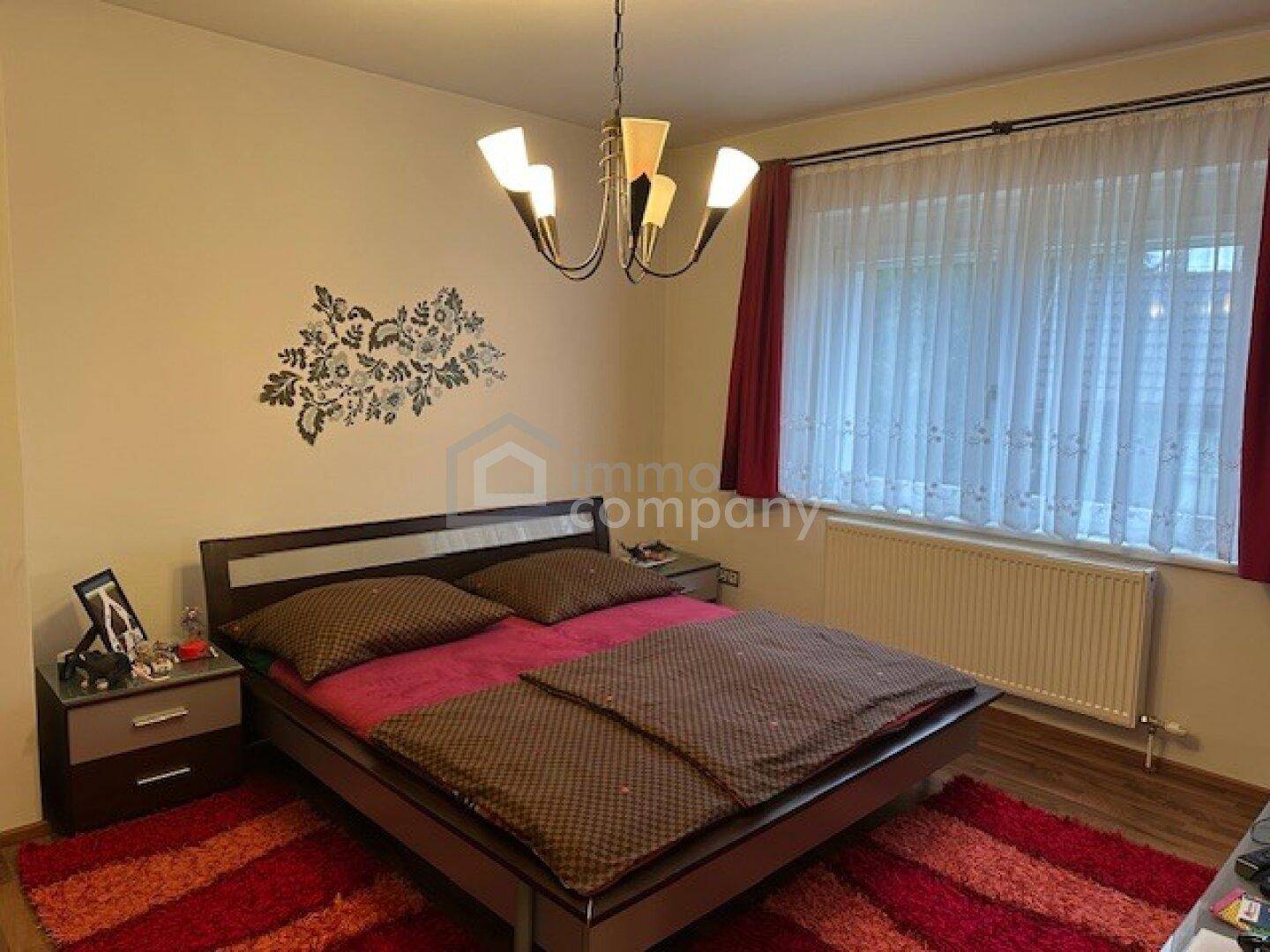 Schlafzimmer mit Doppelbett, rotem Teppich und Fenster mit Vorhängen.