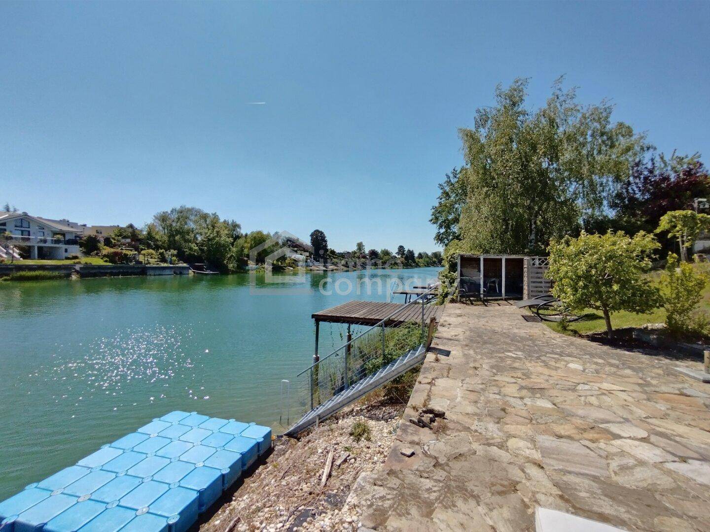 Uferbereich mit Bootsanlegestelle, kleinem Schuppen und schwimmender Plattform am ruhigen See.