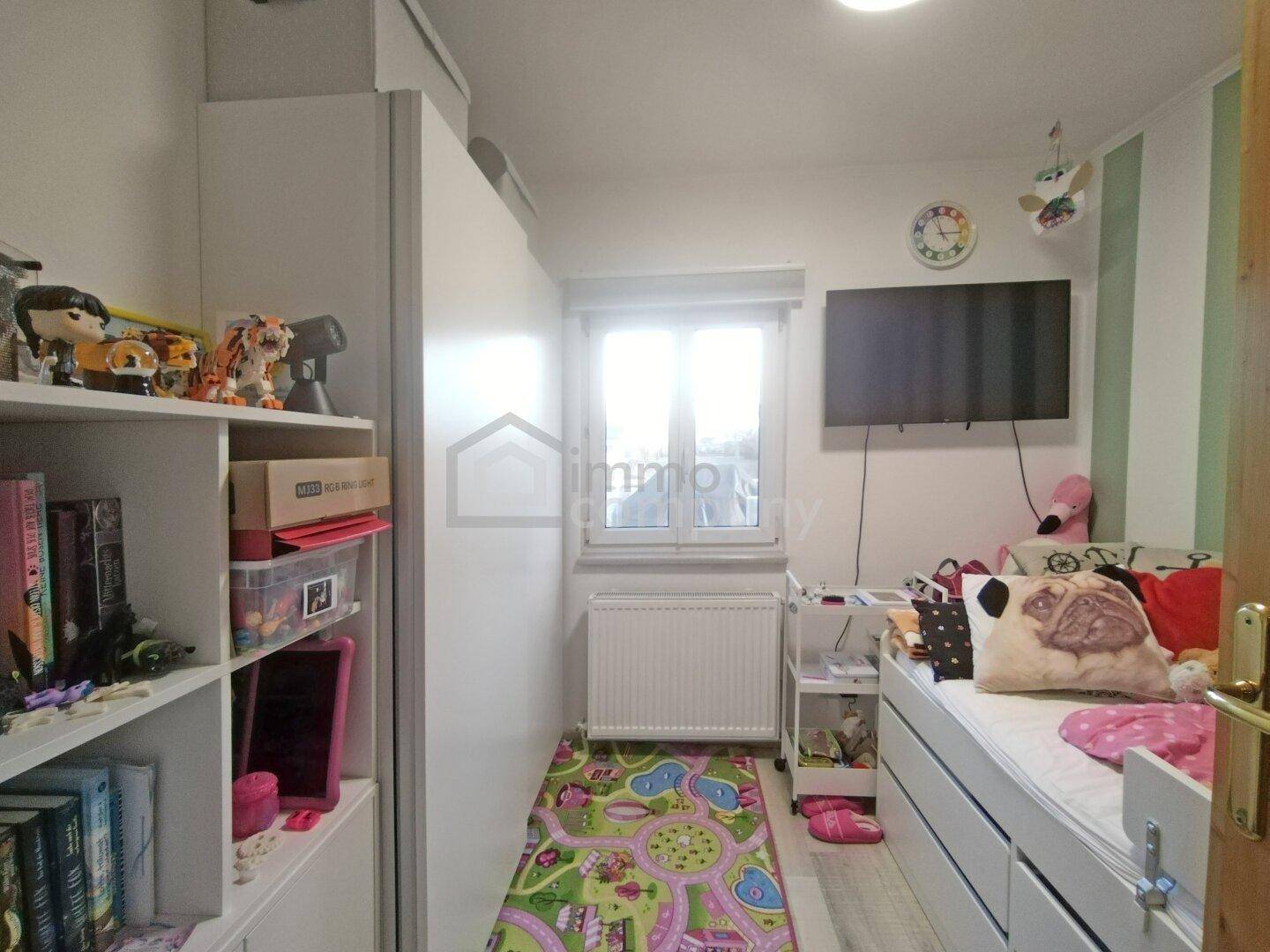 Kinderzimmer mit Bett, Regalen und einem Fenster, hell und freundlich eingerichtet.