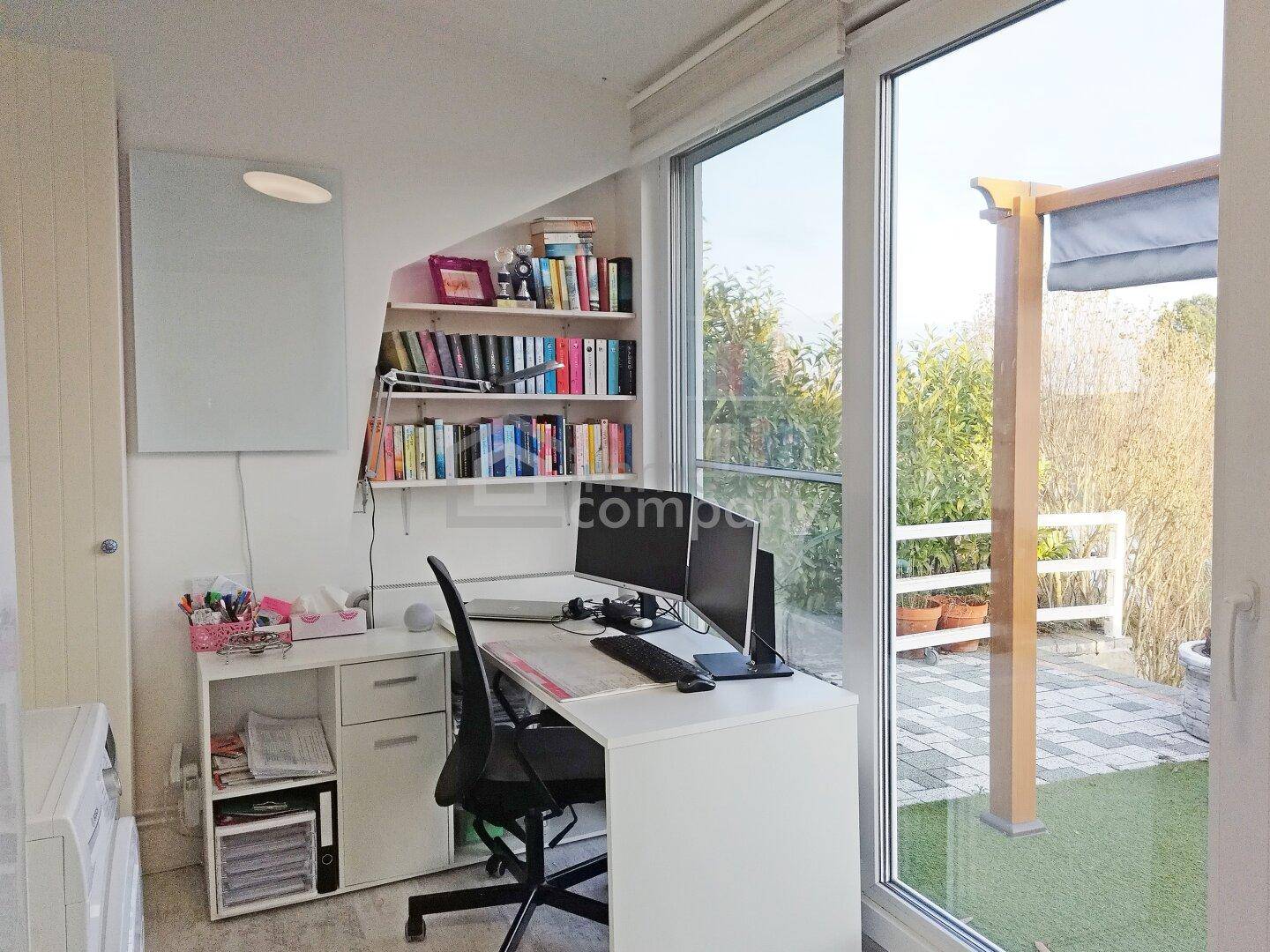 Helles Arbeitszimmer mit Schreibtisch, Bücherregal und Zugang zur Terrasse durch große Fenster.