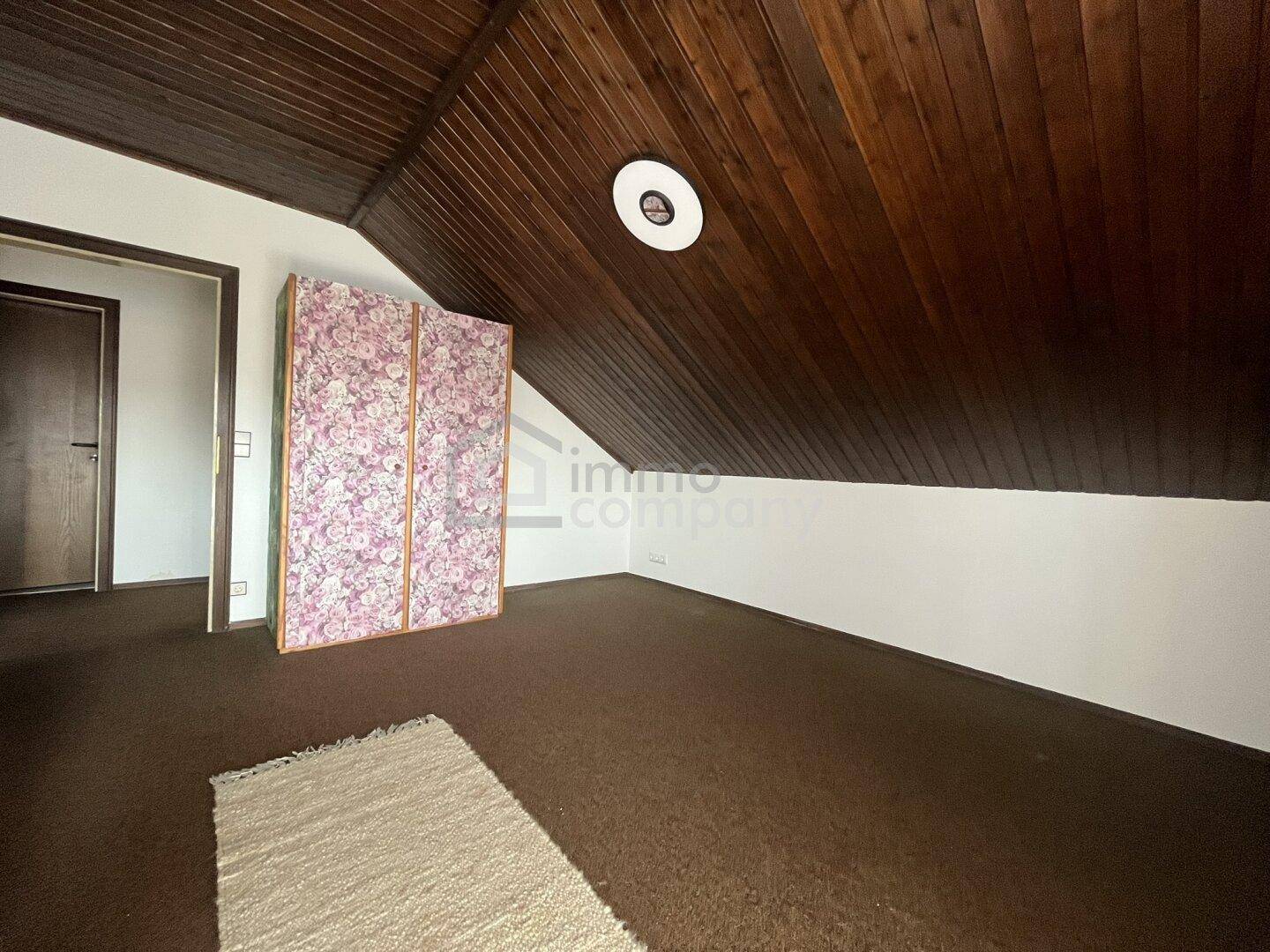 Dachzimmer mit schräger Holztäfelung und Teppichboden, ideal als Gästezimmer.