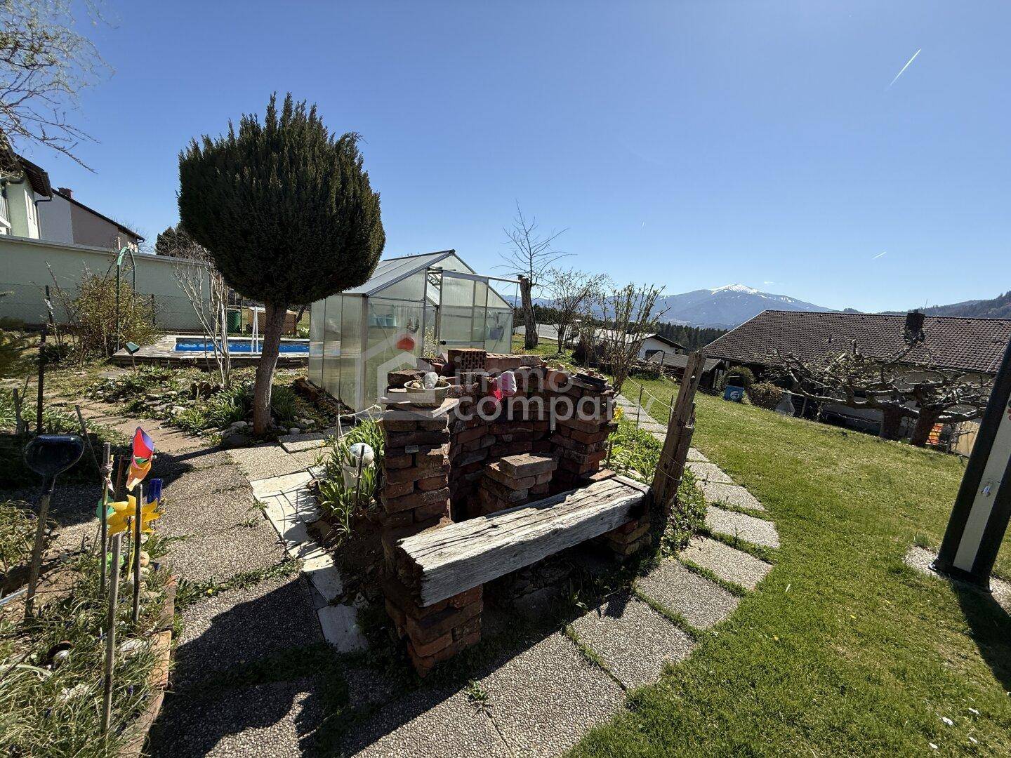 Großer Garten mit gemauerter Sitzbank, Gewächshaus und Pool im Hintergrund.