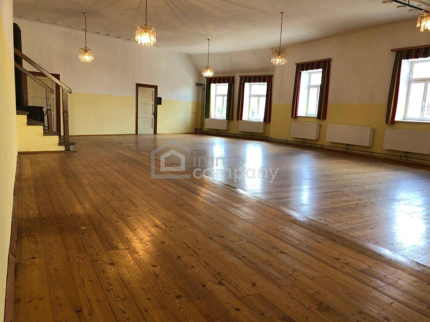 Großer, heller Saal mit Holzdielenboden, hohen Decken und mehreren Kronleuchtern.