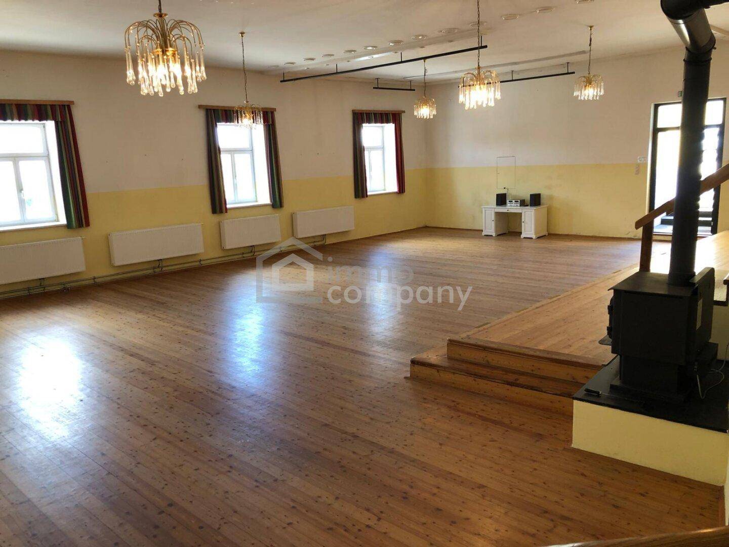 Geräumiger Saal mit Holzdielenboden, Kaminofen und Blick auf die Fensterfront.